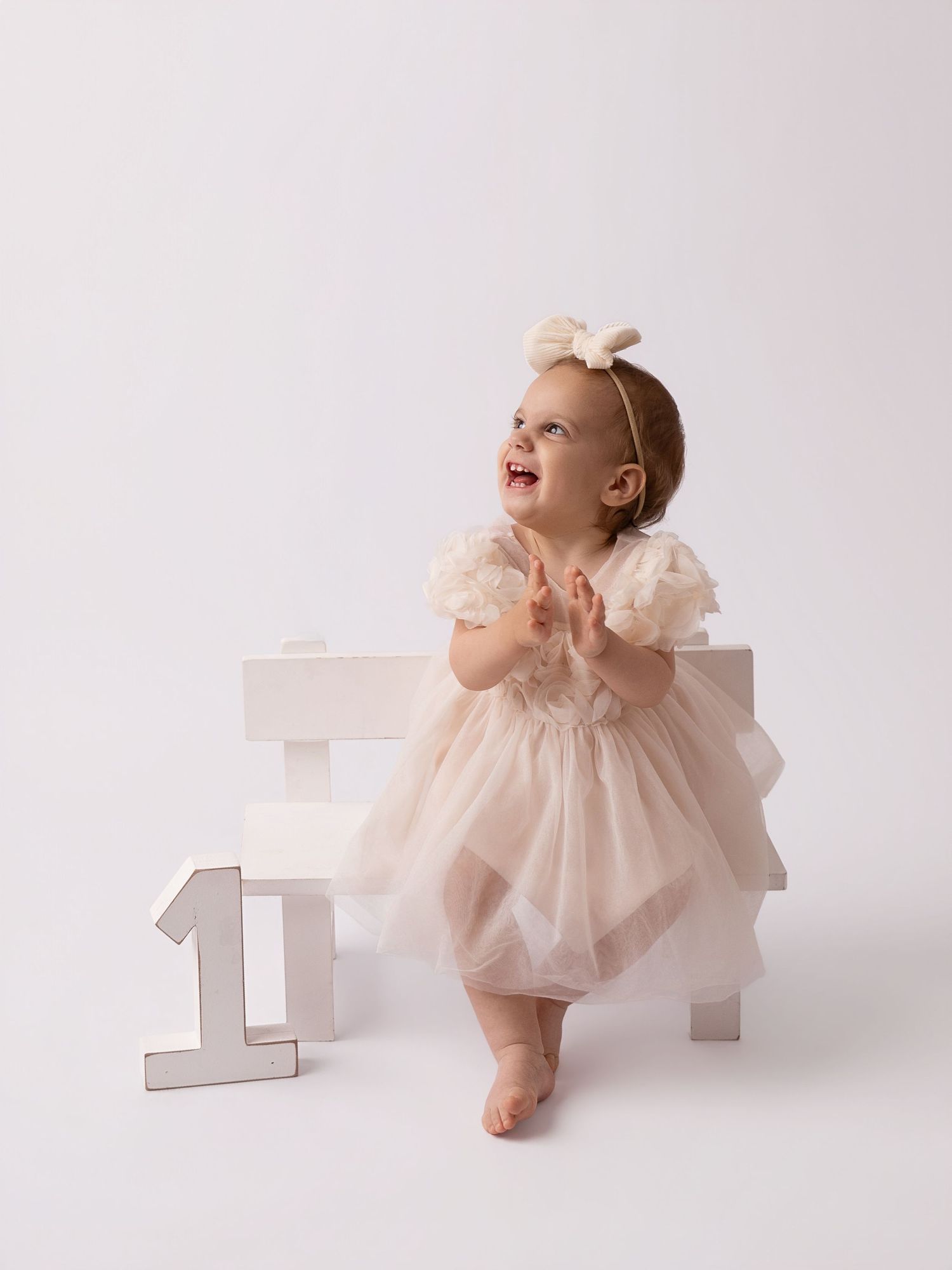 A joyful baby girl in a cream tulle dress celebrates her first birthday beside a white number one.