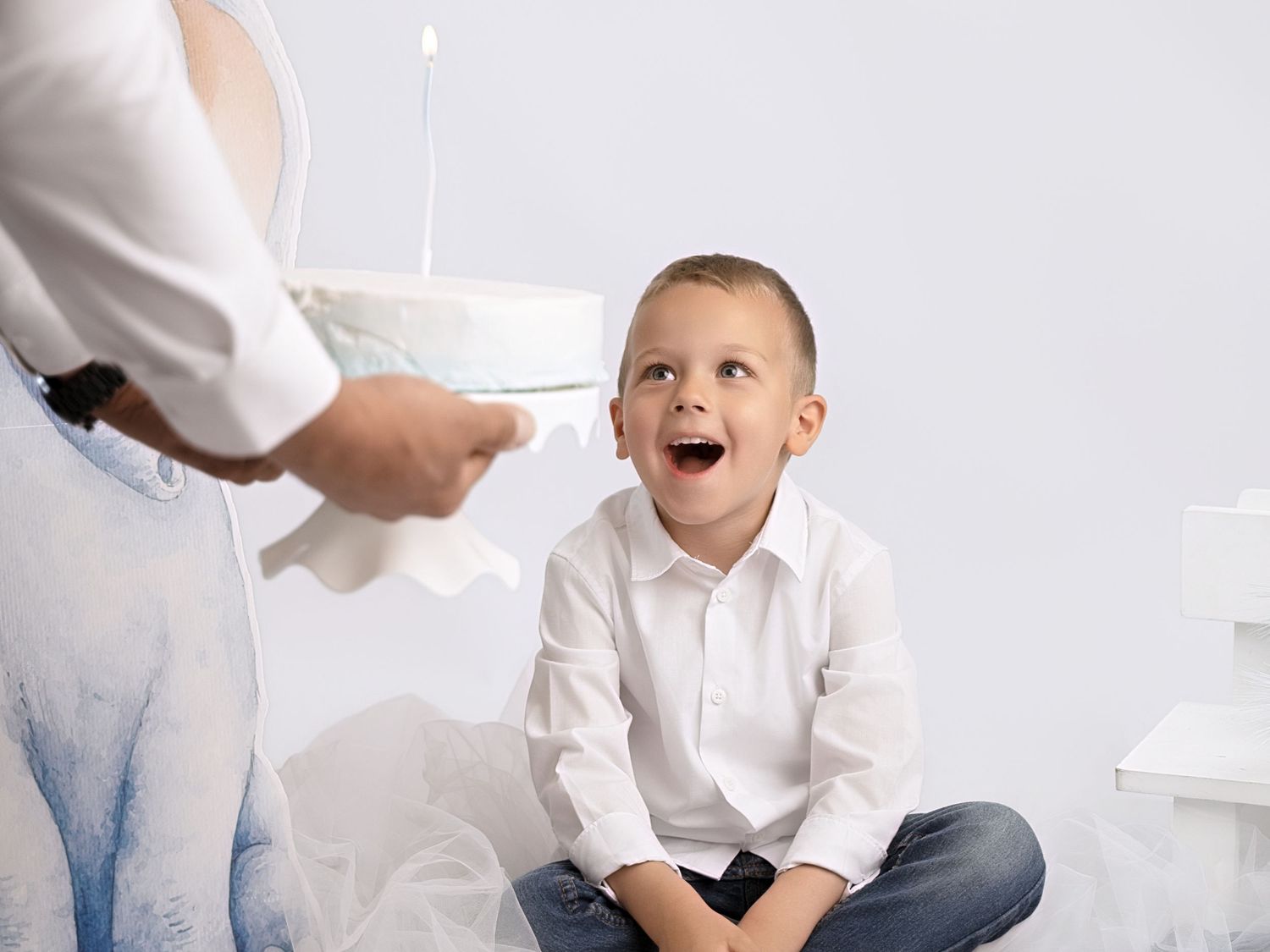 Excited young boy in white shirt reacts with amazement as adult presents a birthday cake with candles.