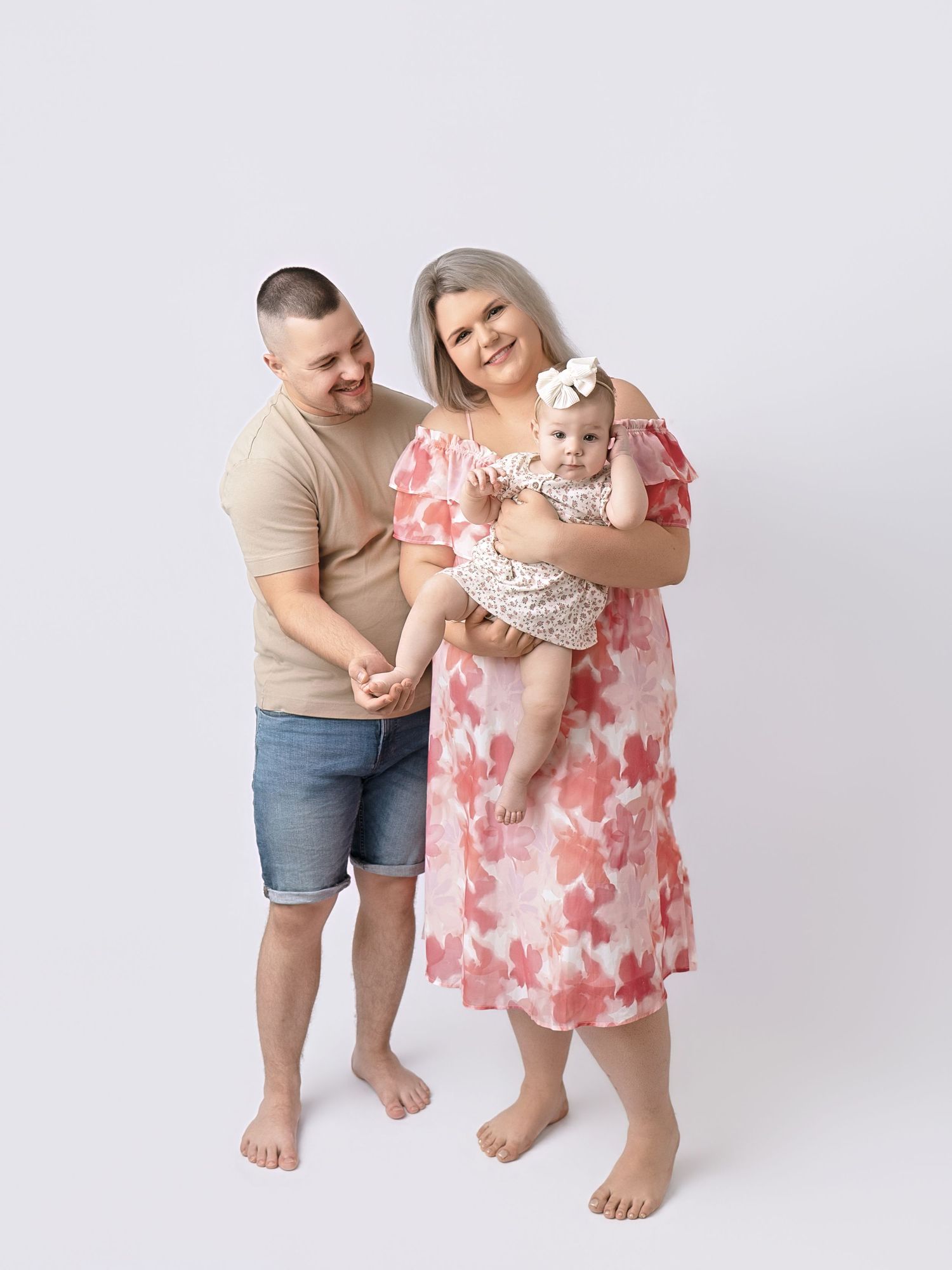 A smiling family with a man in beige, a woman in floral dress, and their newborn baby pose in a studio.