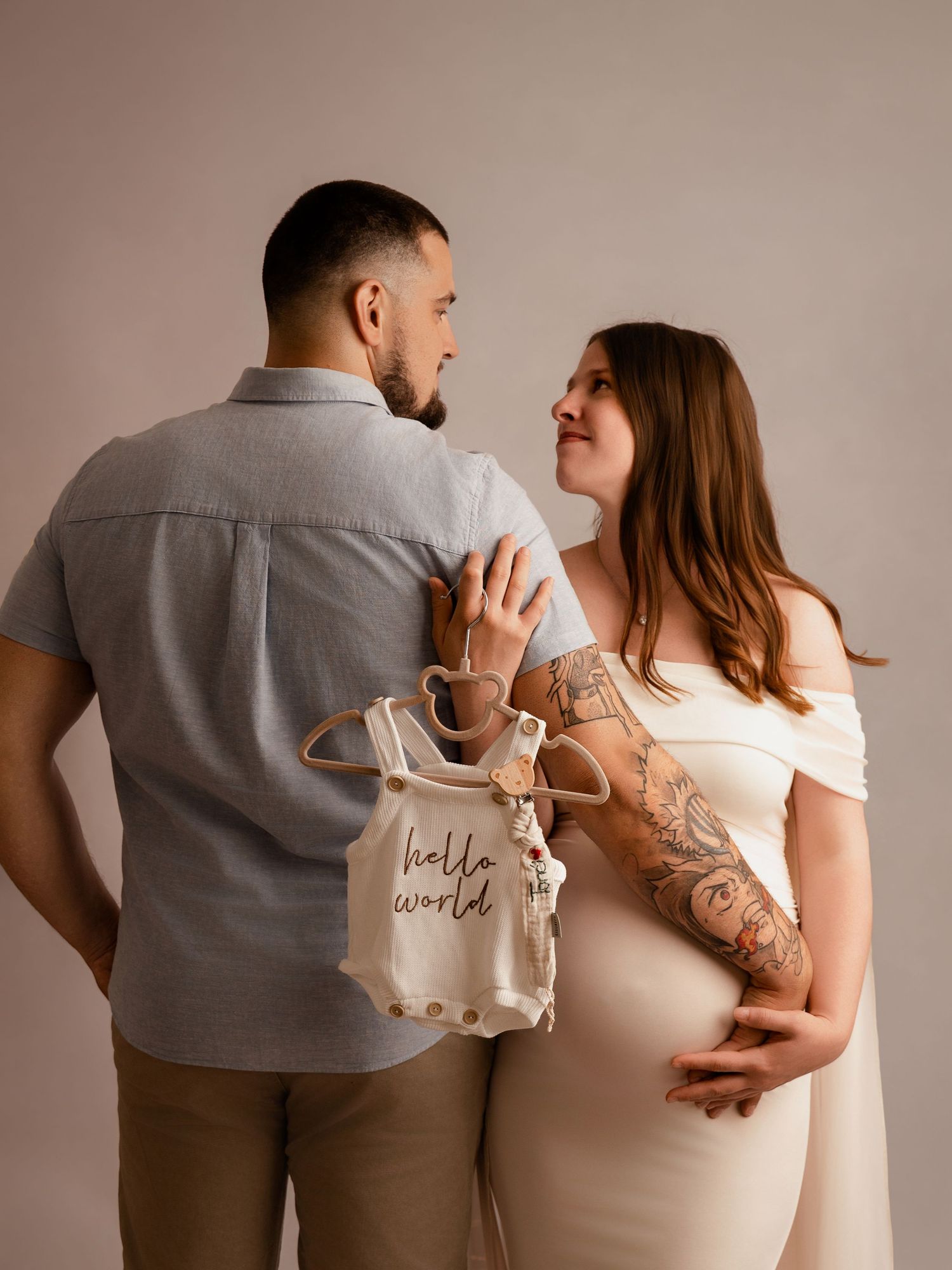 Expecting couple gazing at each other while holding a tiny hello world baby onesie in a studio setting.