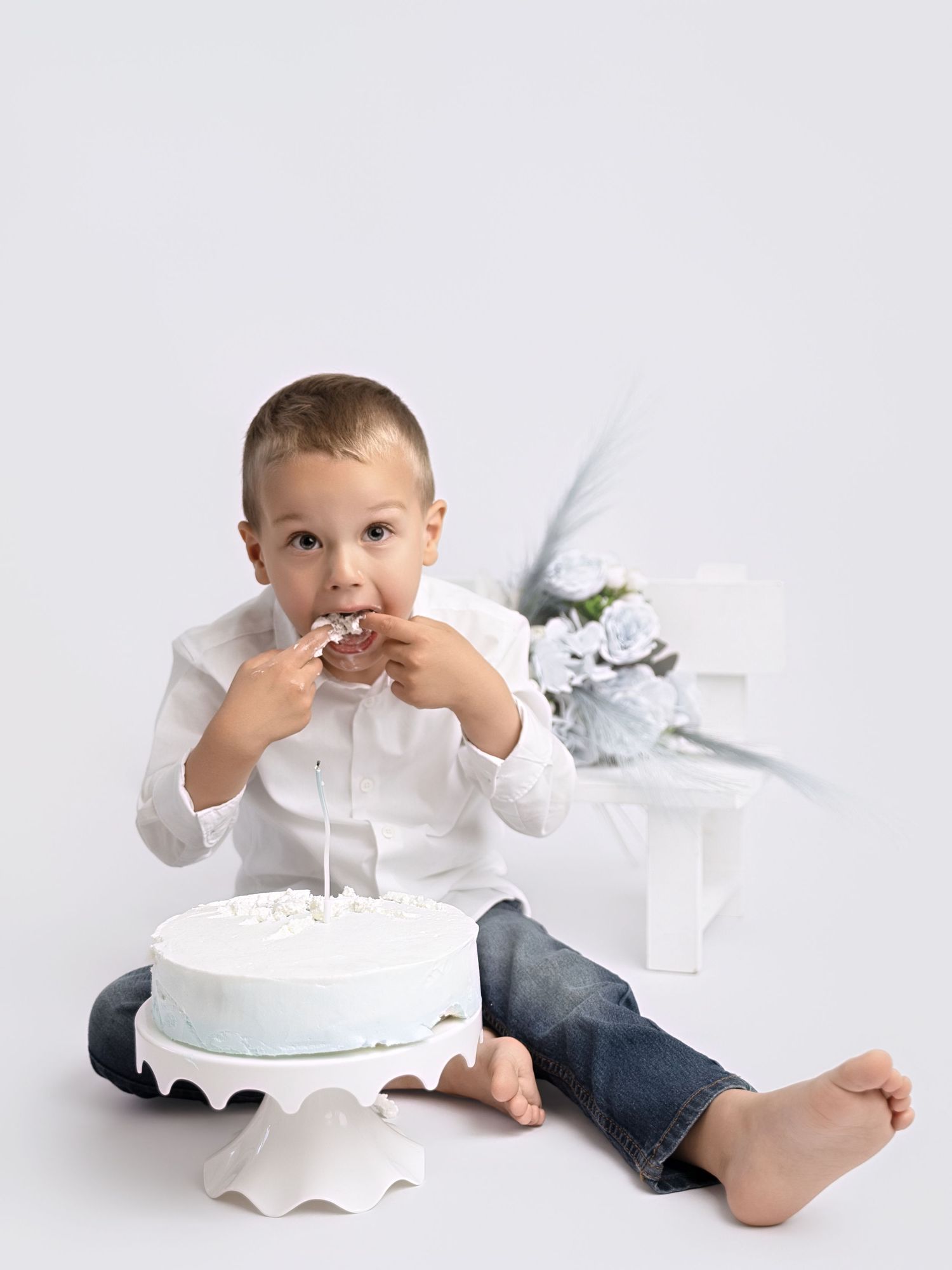 Young boy in white shirt and jeans eating birthday cake during a smash cake photoshoot on white background.