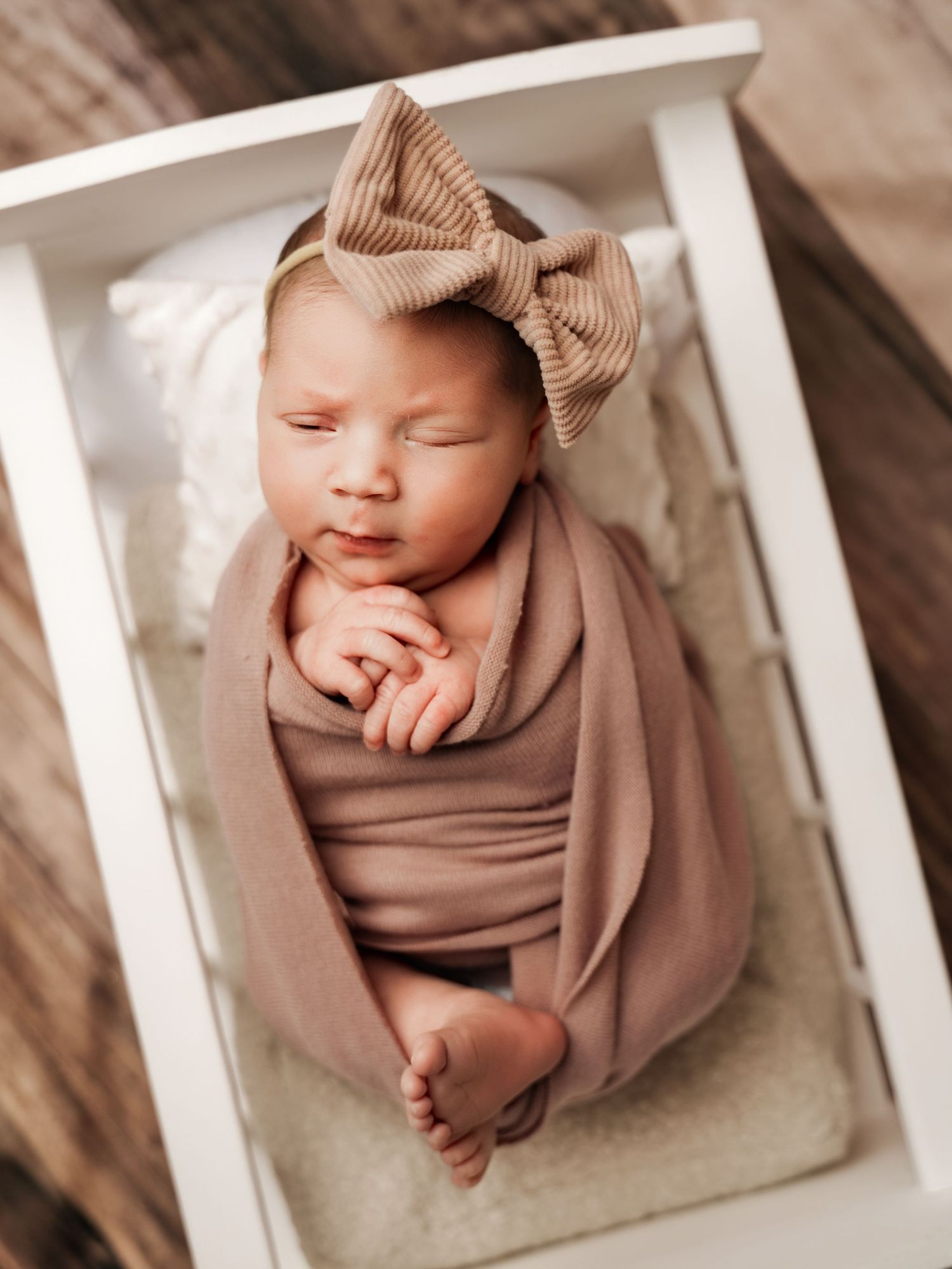 Sleeping newborn baby wrapped in a soft taupe swaddle with a bow headband, resting in a white mini crib.