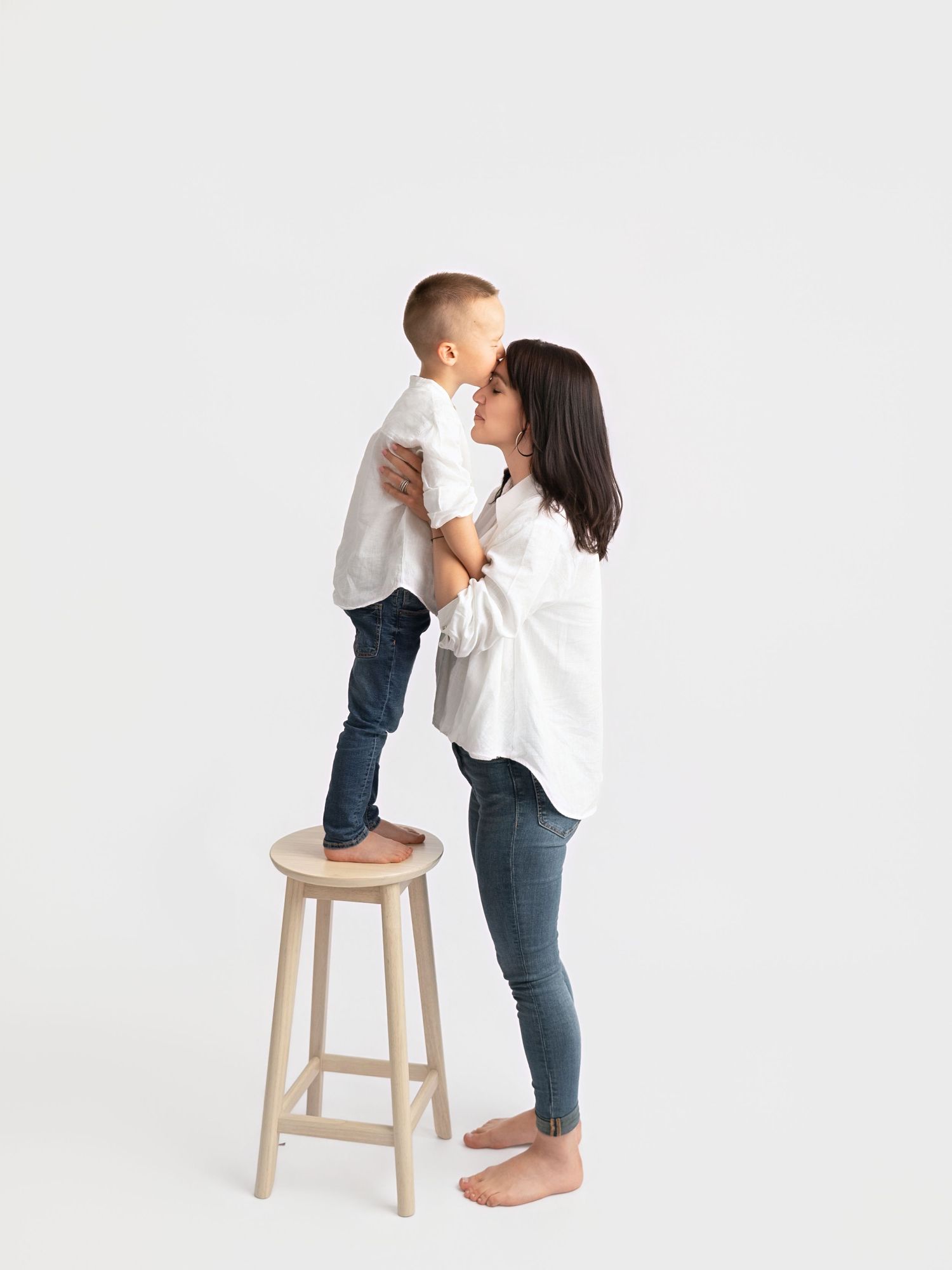 A mother in a white shirt and jeans kisses her young son standing on a wooden stool against a white background.