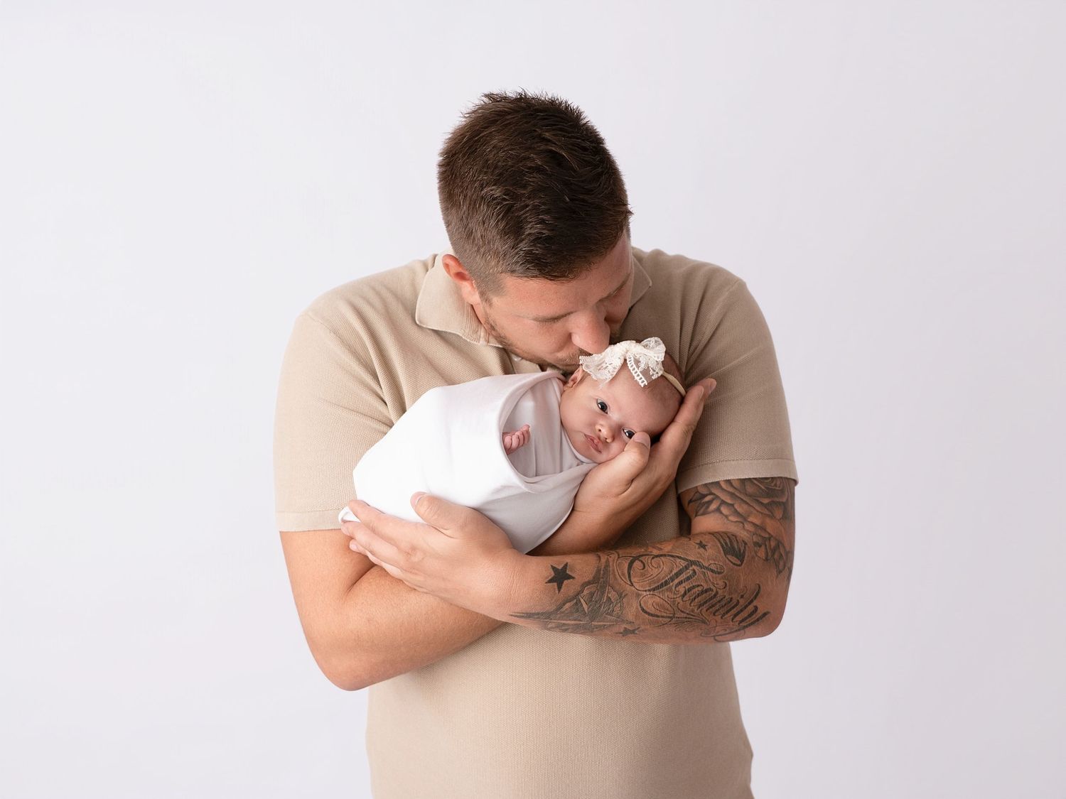 Father in beige polo with tattoos kissing swaddled newborn baby against white background.