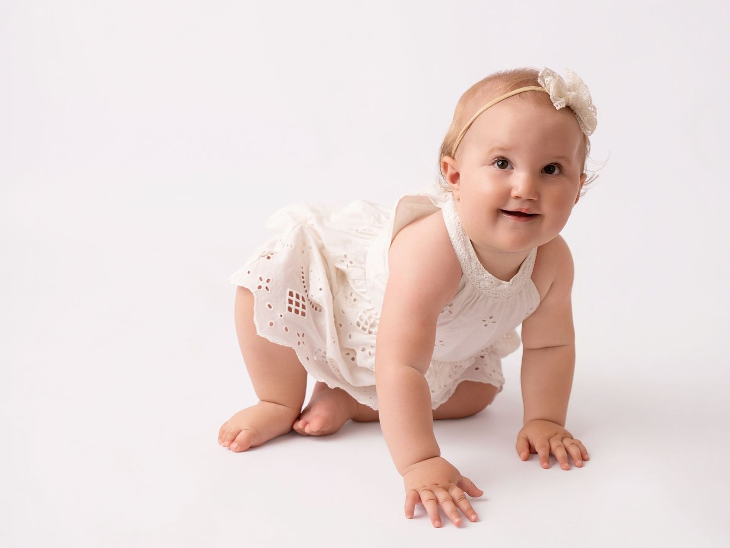Adorable baby girl in white lace romper crawling on white background, wearing a floral headband.