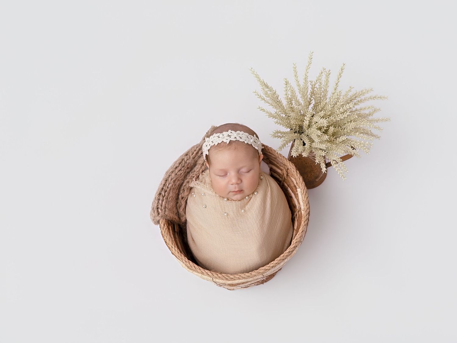 Newborn baby sleeping peacefully in a wicker basket, wrapped in beige, wearing a lace headband, beside a small plant.