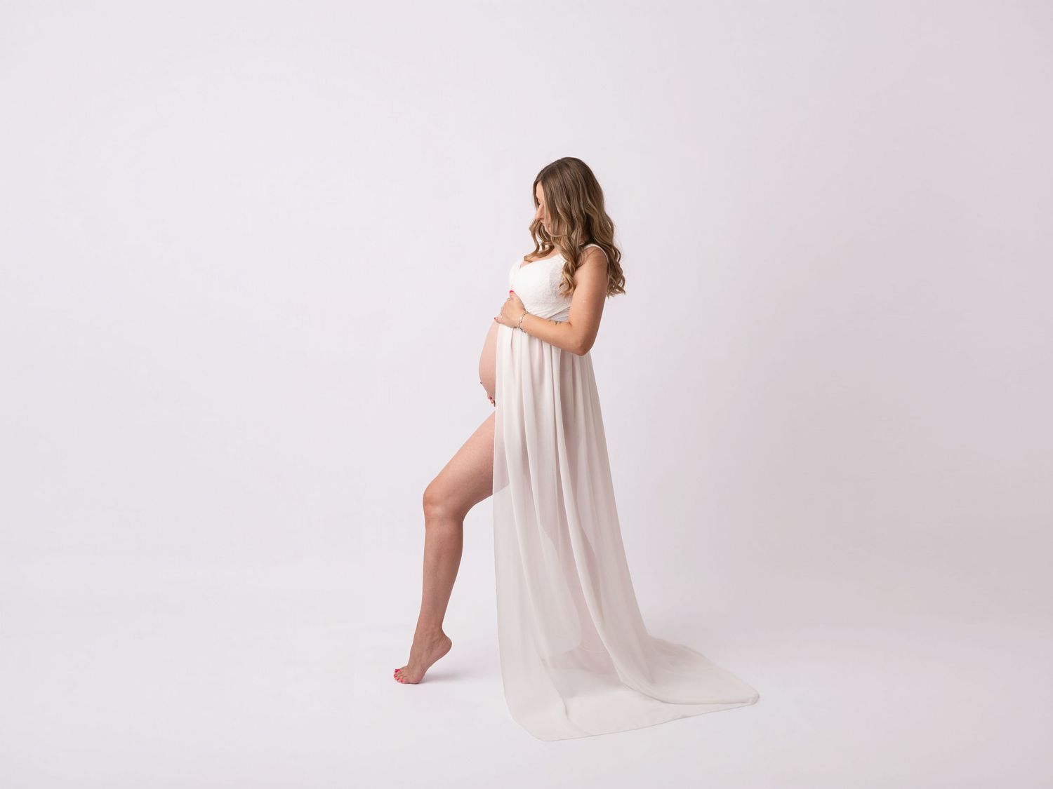 Elegant pregnant woman in flowing white maternity gown posing gracefully against white background.