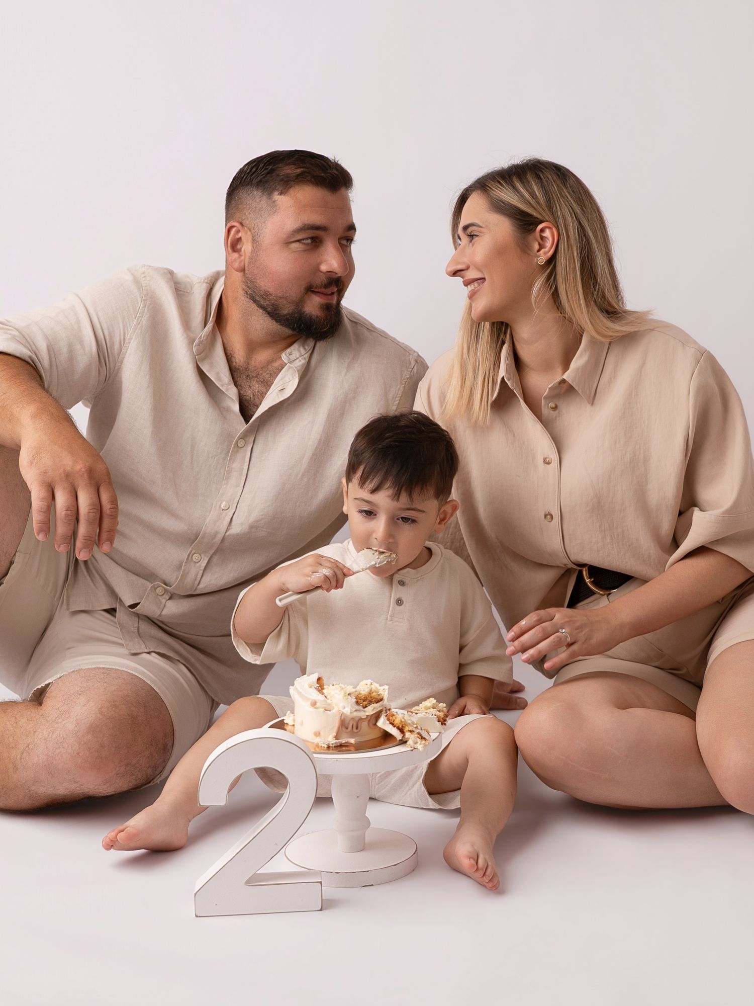 Family of three in matching beige outfits celebrating toddler's 2nd birthday with cake smash photo session.