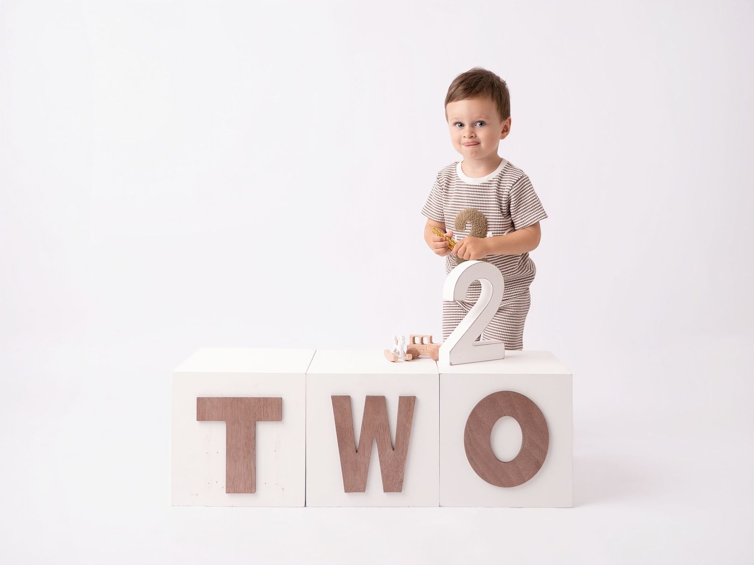 Toddler boy standing behind white blocks spelling TWO, celebrating his 2nd birthday in a beige outfit.