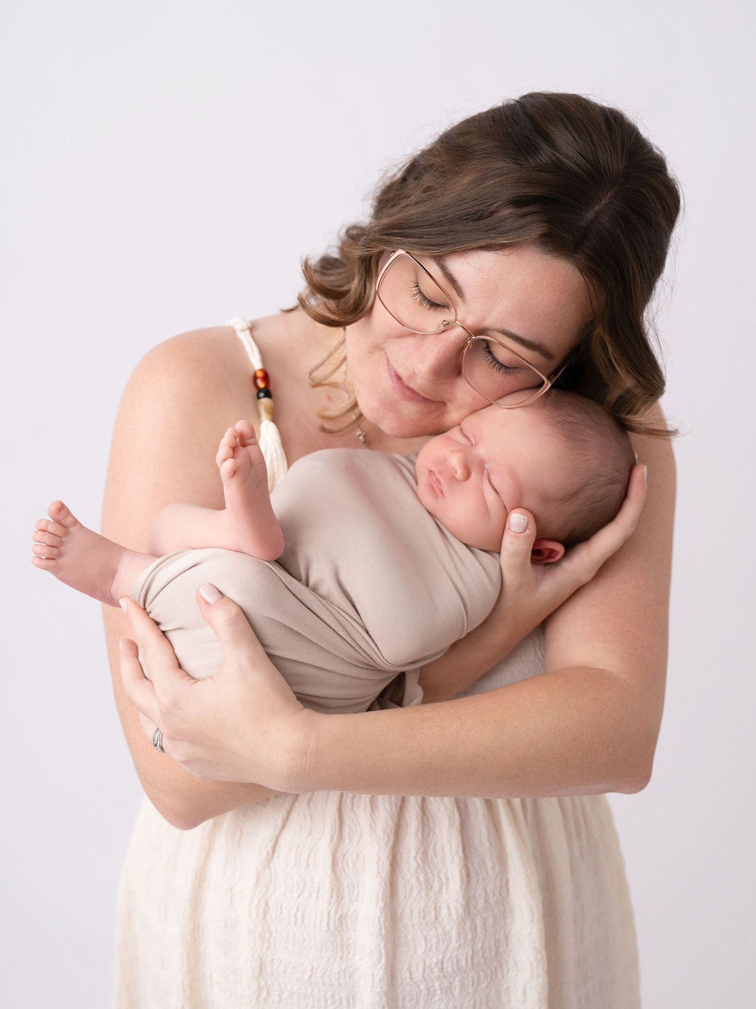 A mother with glasses tenderly holds her sleeping newborn baby wrapped in a beige swaddle against a white background.