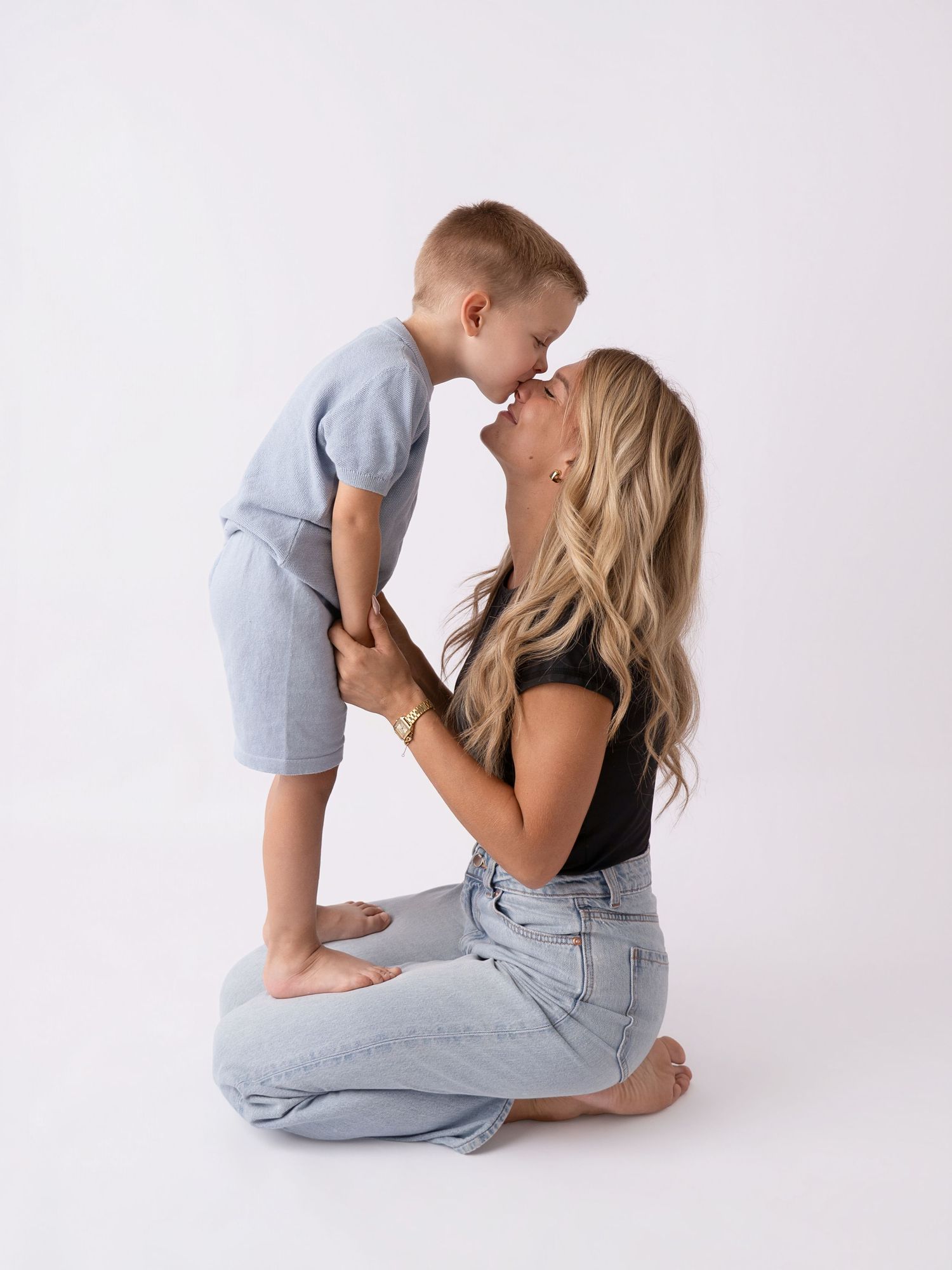 A blonde woman sits on the floor sharing a sweet nose-to-nose moment with a young boy in a light blue outfit.