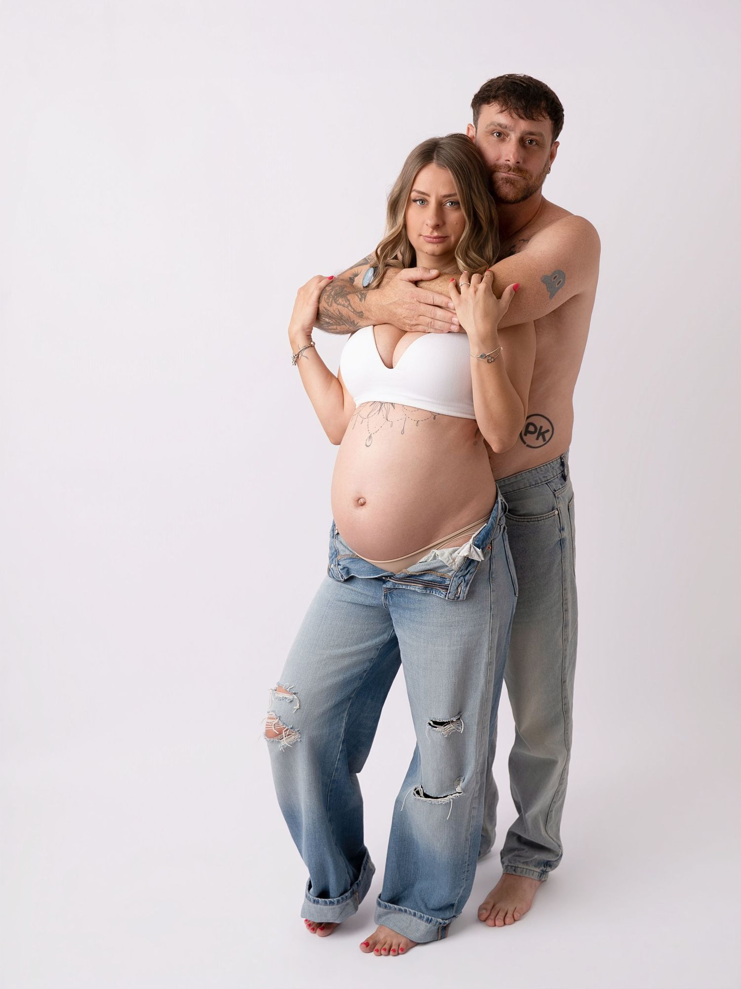 Pregnant couple posing together, woman in white crop top and ripped jeans, man shirtless with tattoos, maternity photo.