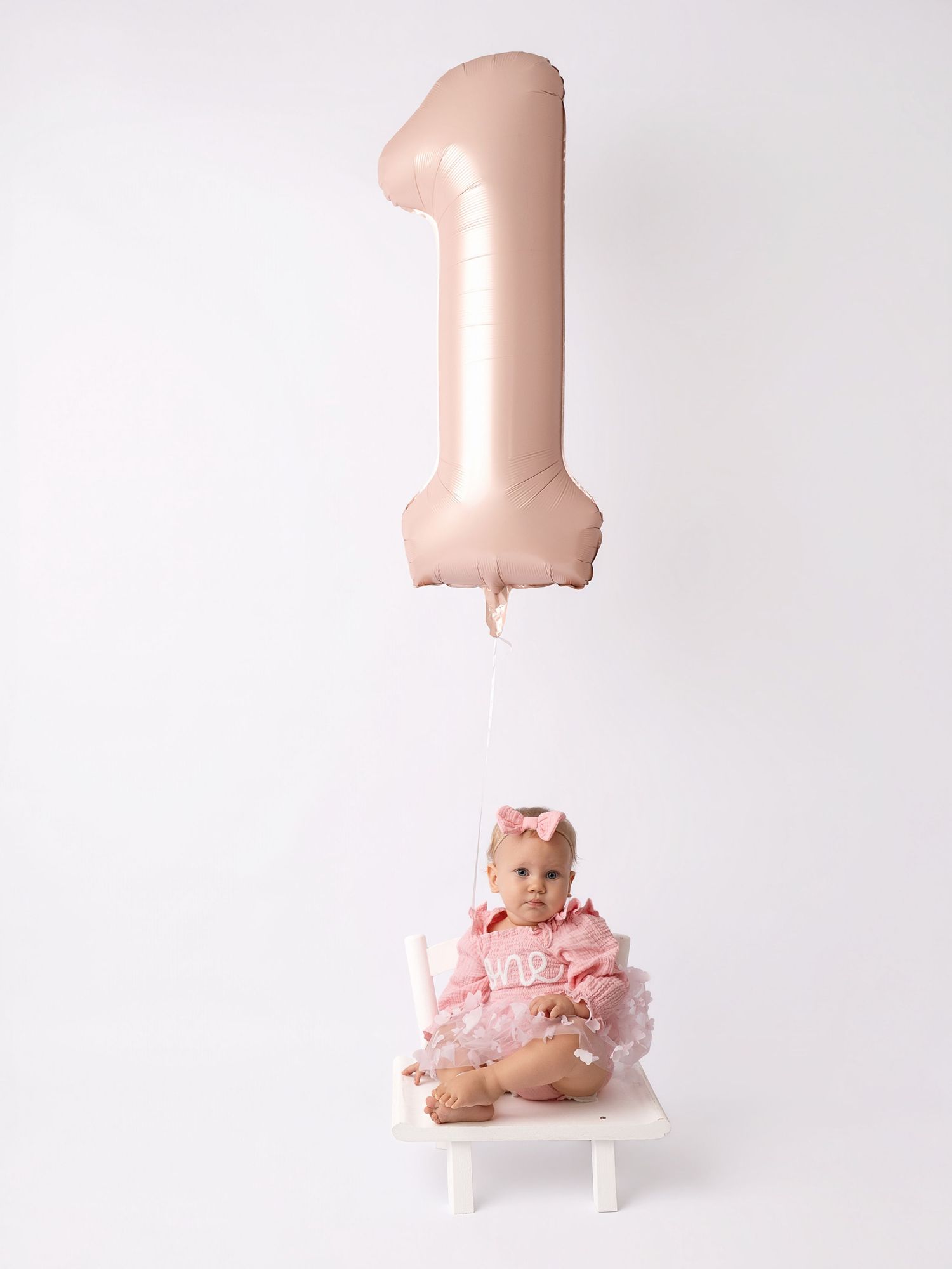 Baby girl in pink outfit sits under a rose gold number 1 balloon for her first birthday photo.