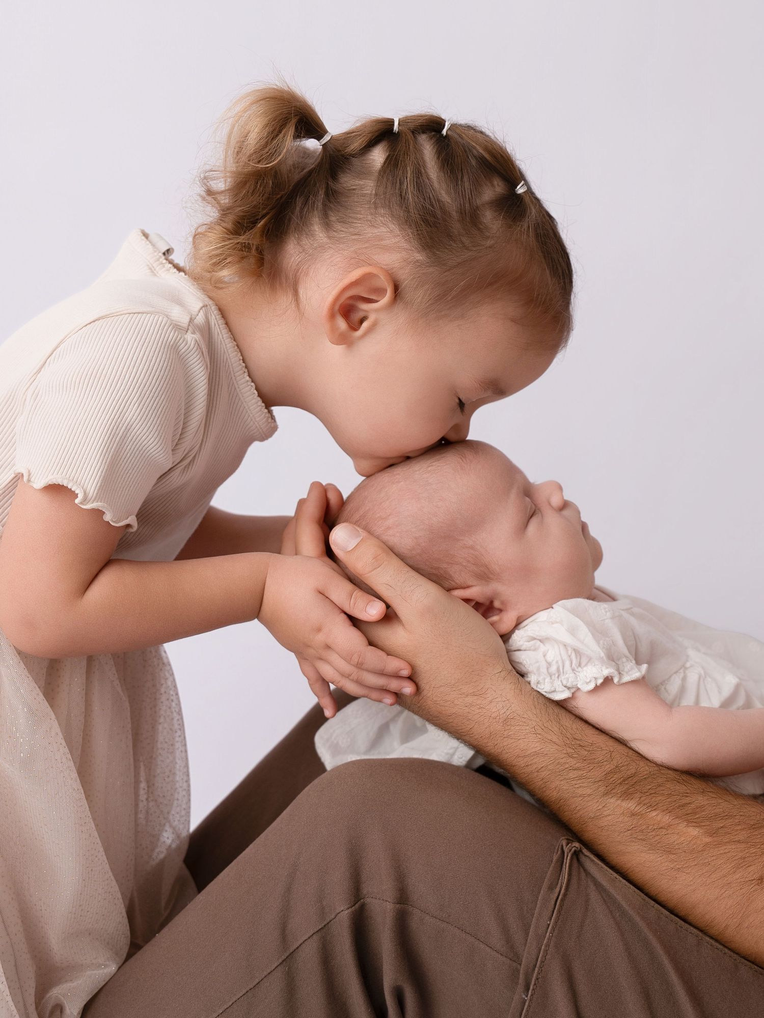 Young girl with pigtails kissing newborn baby held in father's hands against white background.