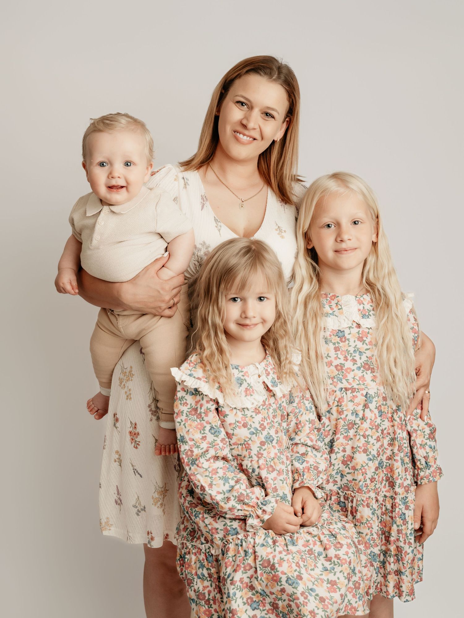 A smiling mother holds a baby while posing with two blonde girls in floral dresses for a studio family portrait.