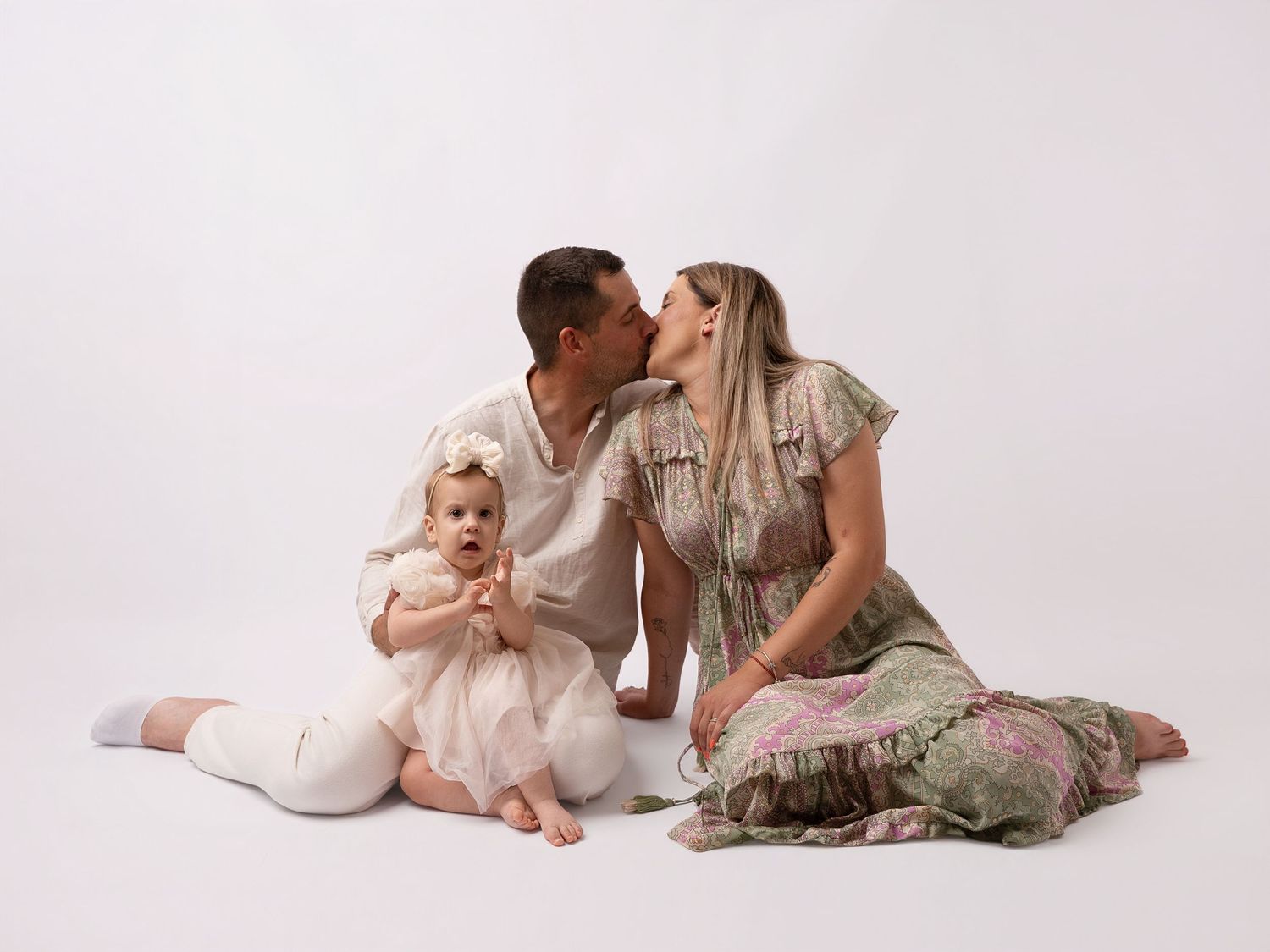 A couple kisses while their baby girl in white sits in front on a studio white background.