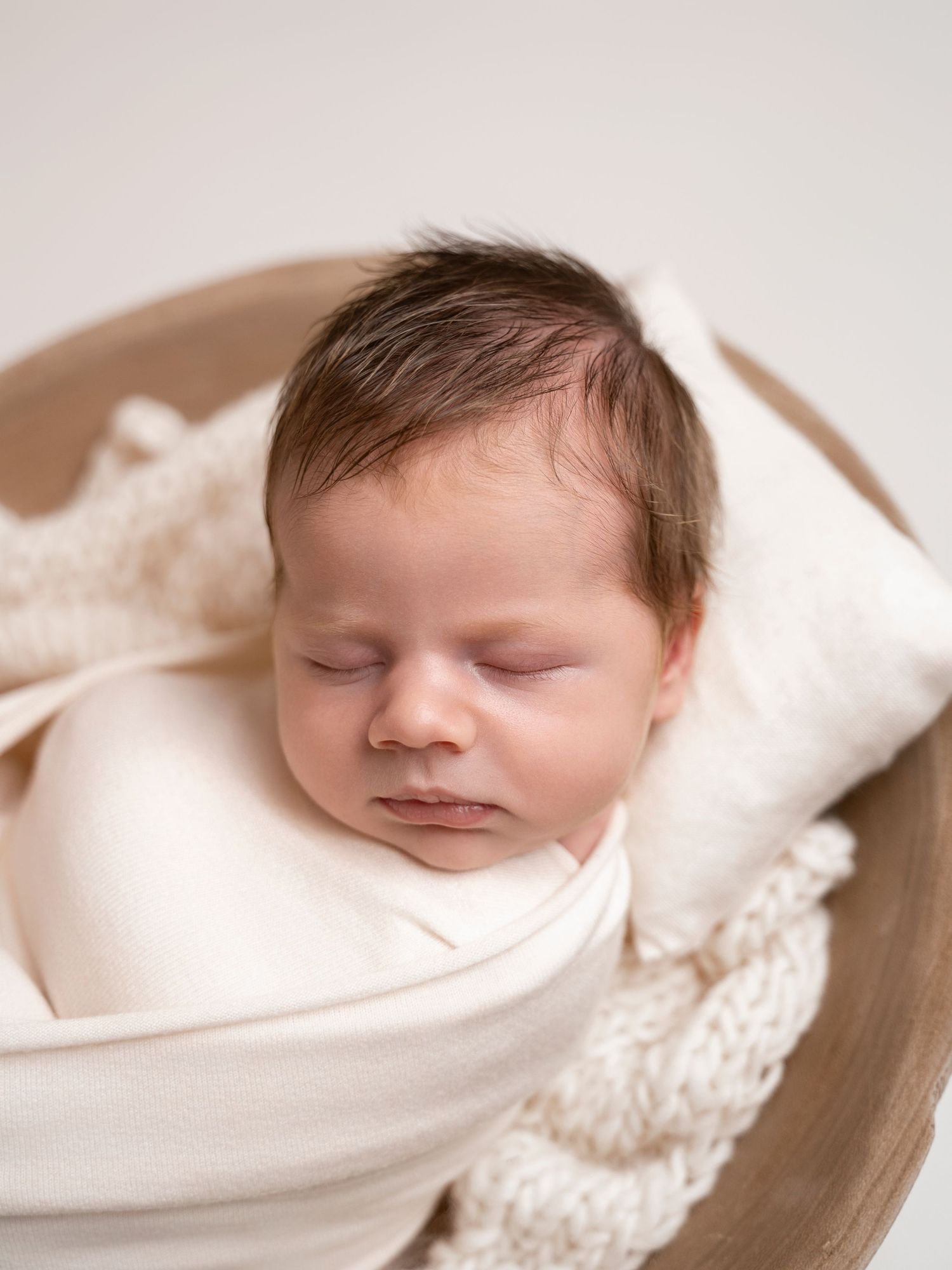 Peaceful newborn baby sleeping wrapped in cream fabric inside a wooden bowl prop.