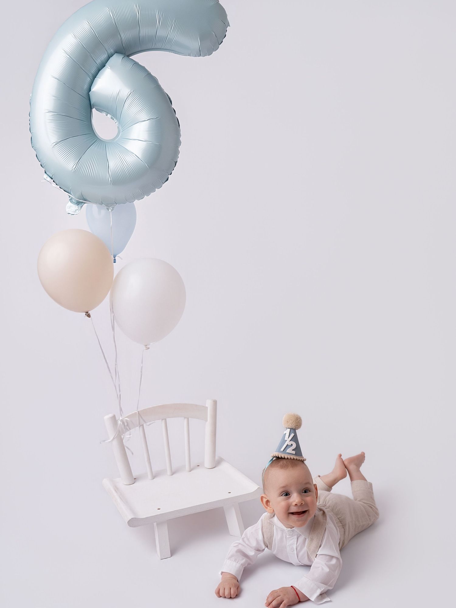Cute baby in party hat lies beside white chair with blue number 6 and pastel balloons for 6-month milestone.