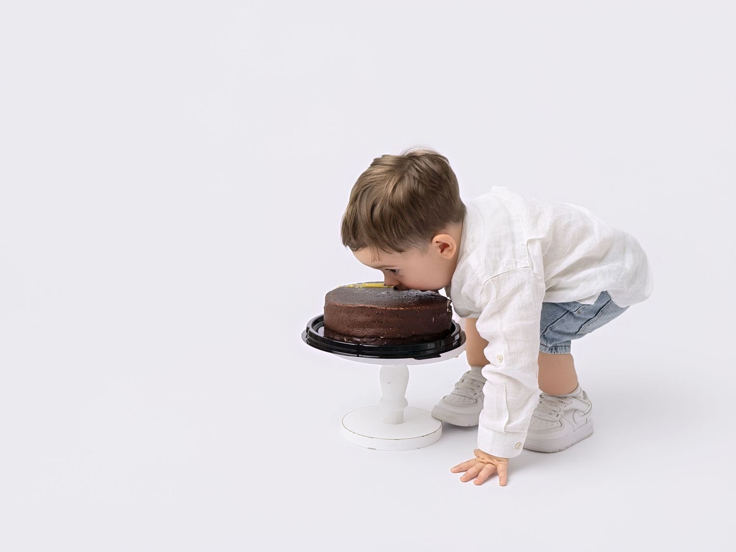 Toddler boy in white shirt and denim shorts leaning over a chocolate cake on a white cake stand.