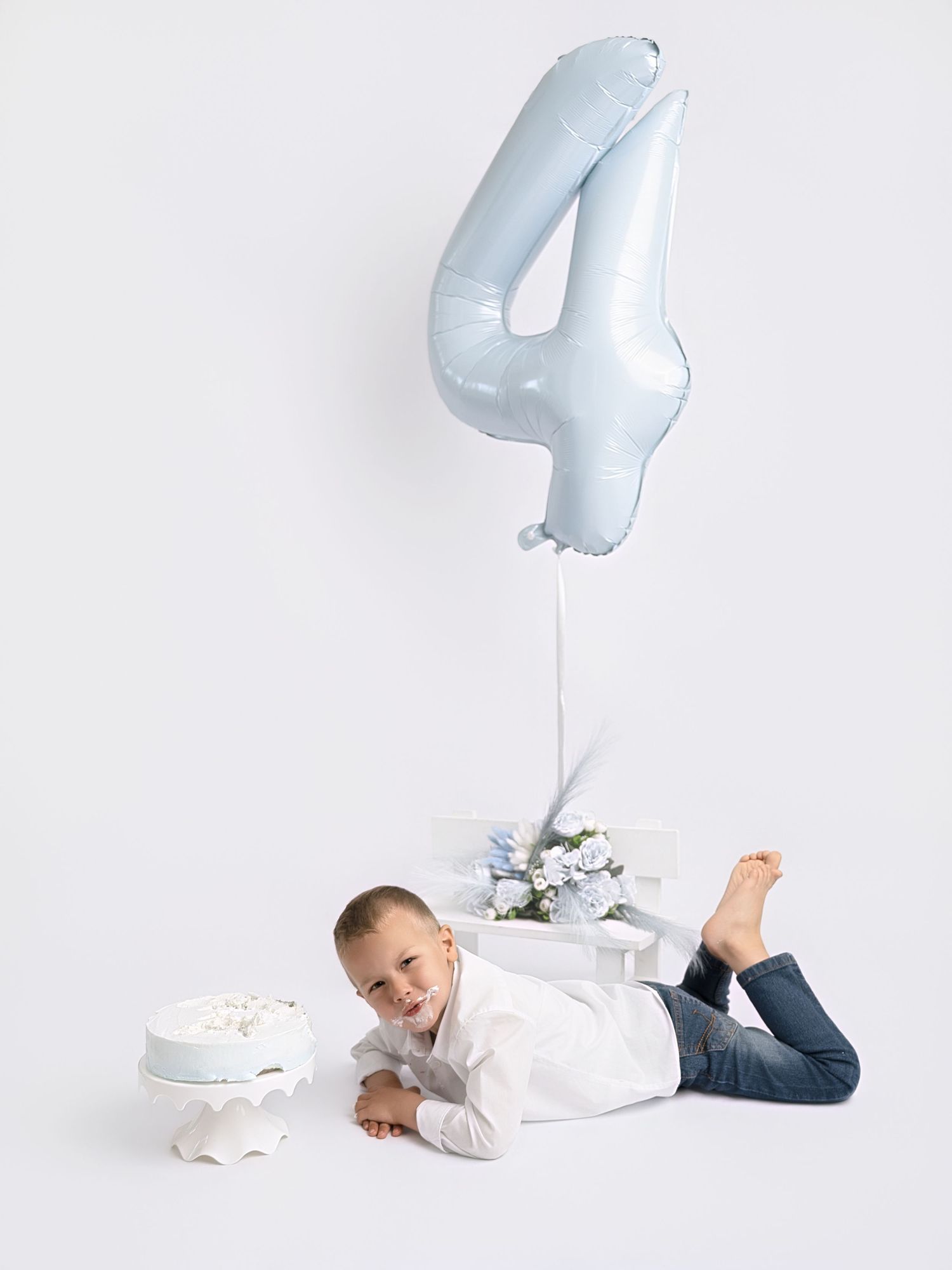 A young boy lying on the floor smiling next to a white cake, with a blue number 4 balloon above him.