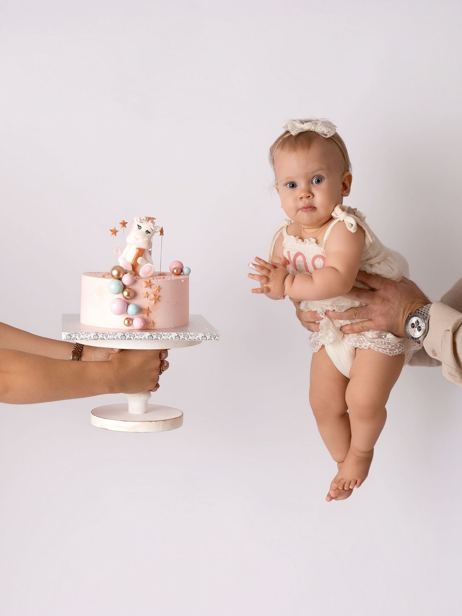 Baby girl in lace outfit held by parent near decorated pink first birthday cake on white background.