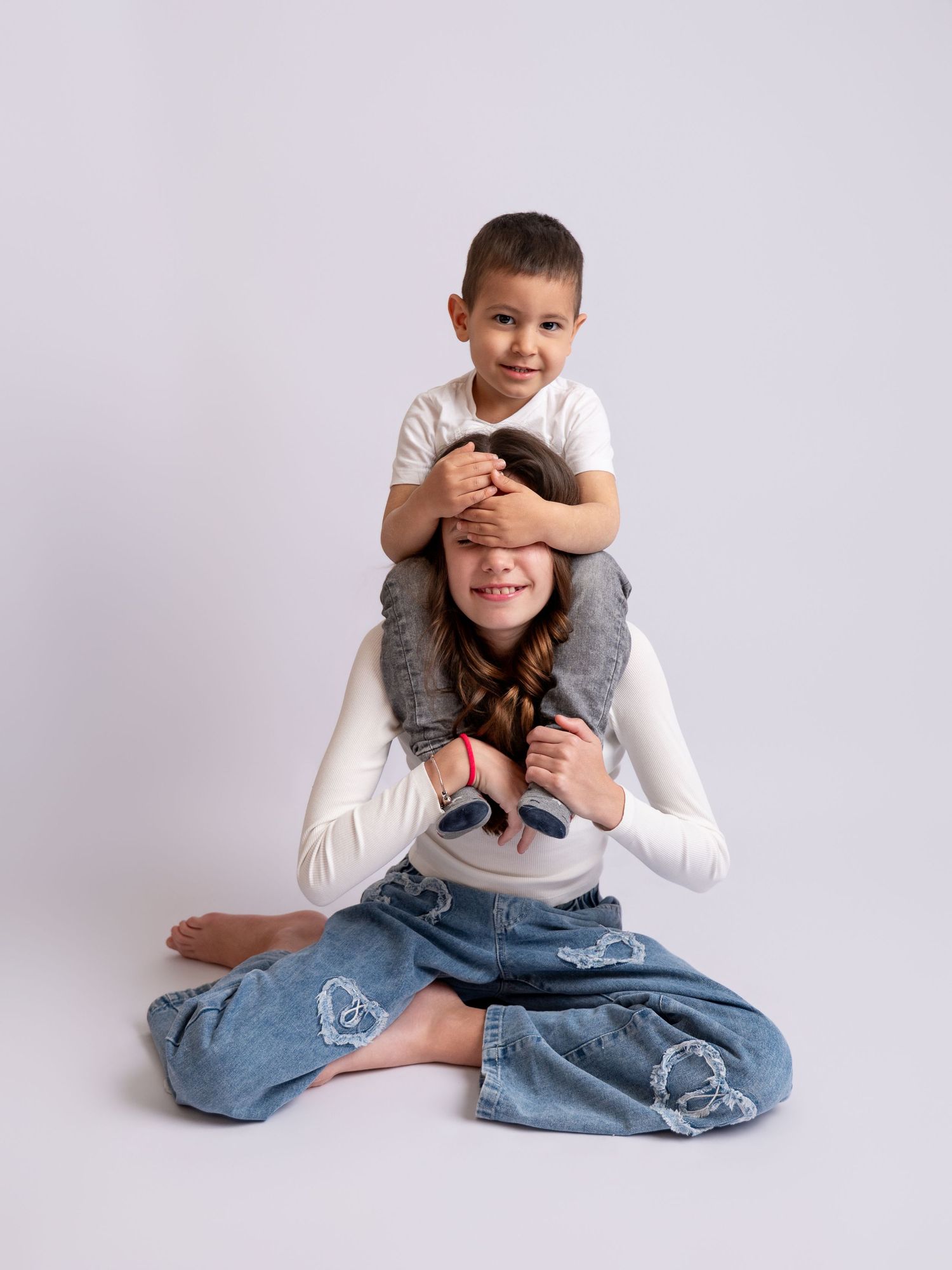 Two siblings pose playfully, with a boy sitting on his sister's shoulders in a white studio.