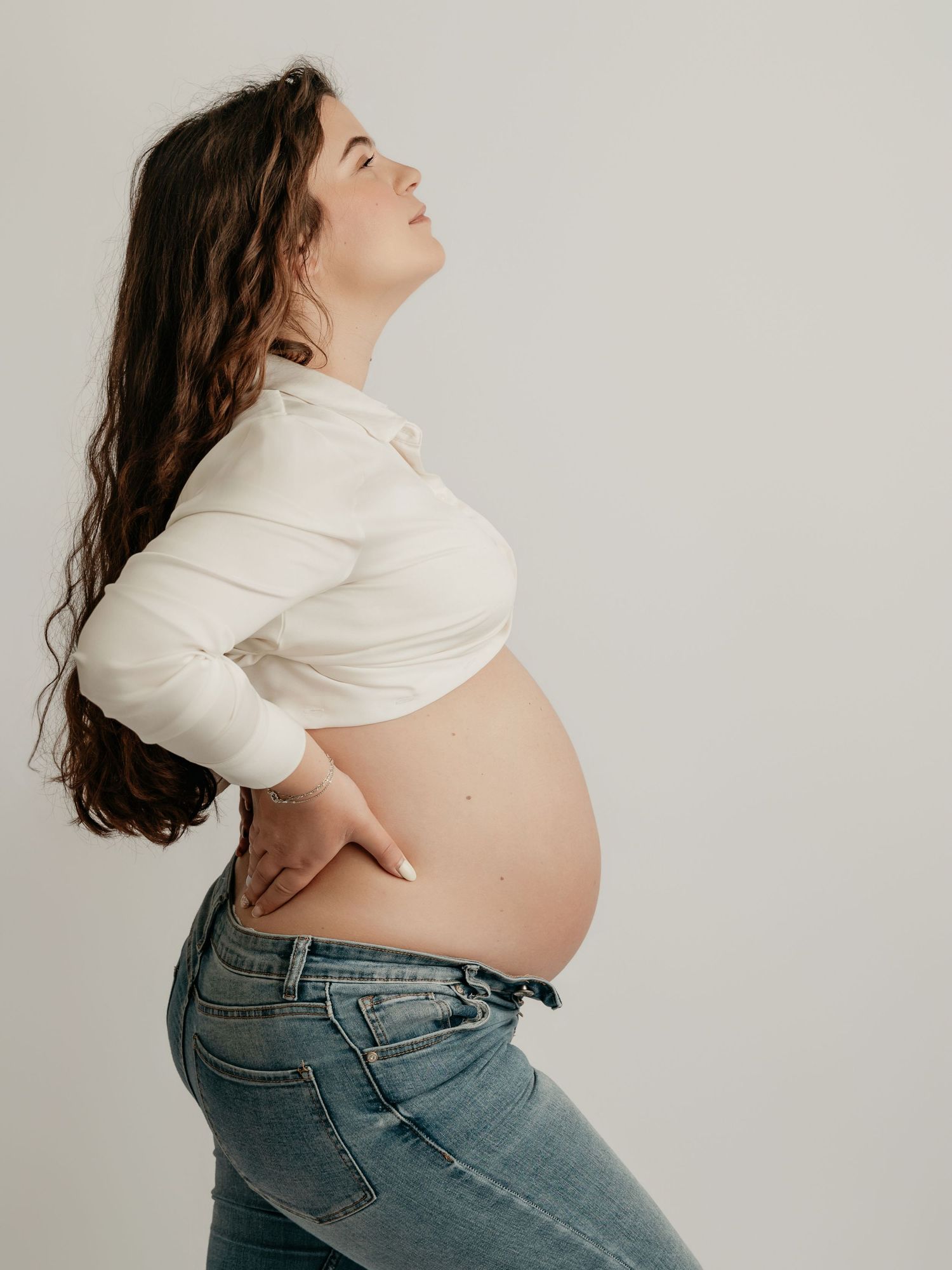 Pregnant woman in white crop top and jeans holding her baby bump, side profile view against white background.