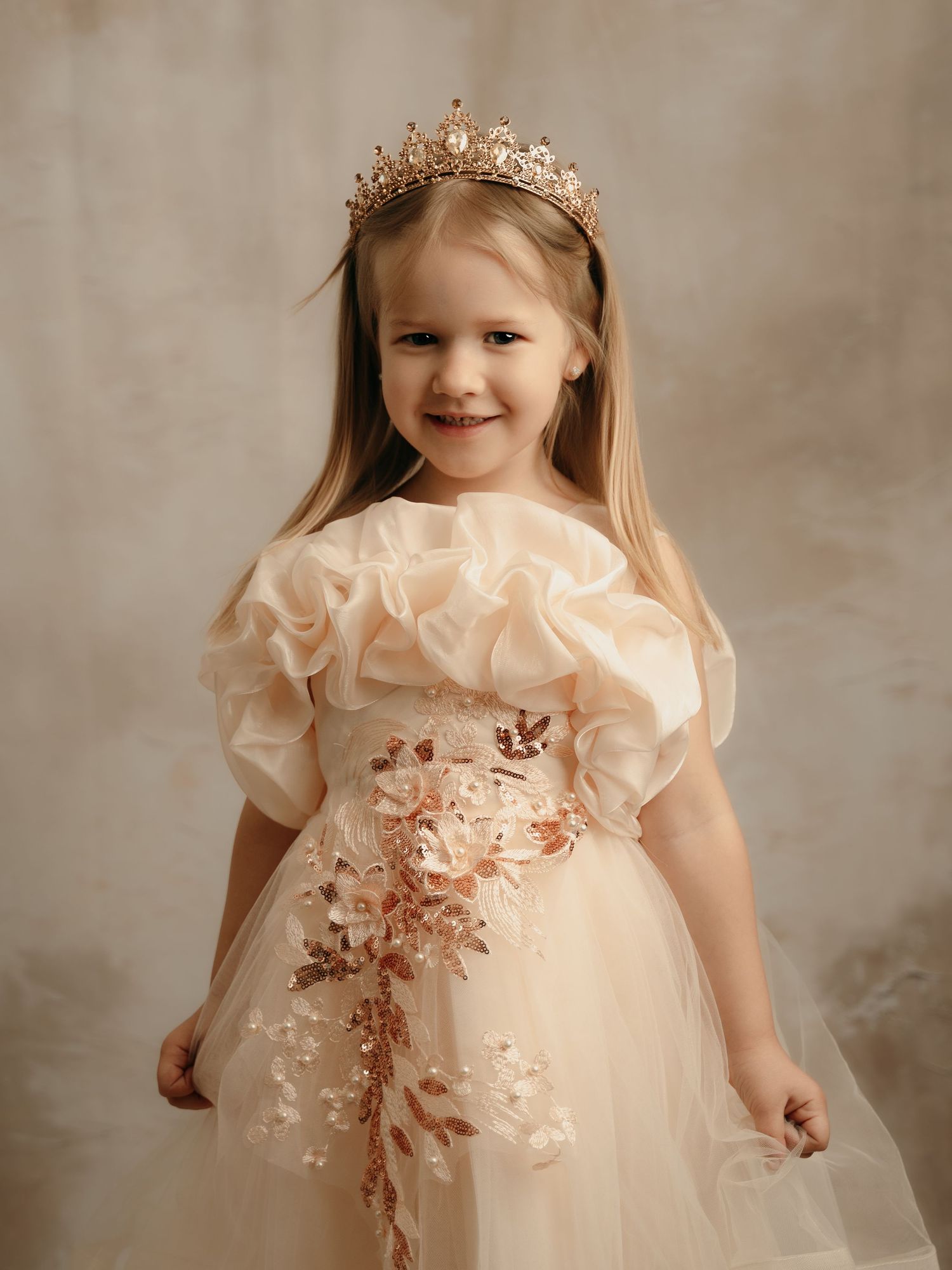Smiling girl in an elegant champagne tulle ball gown with floral sash and gold crown tiara accessory.