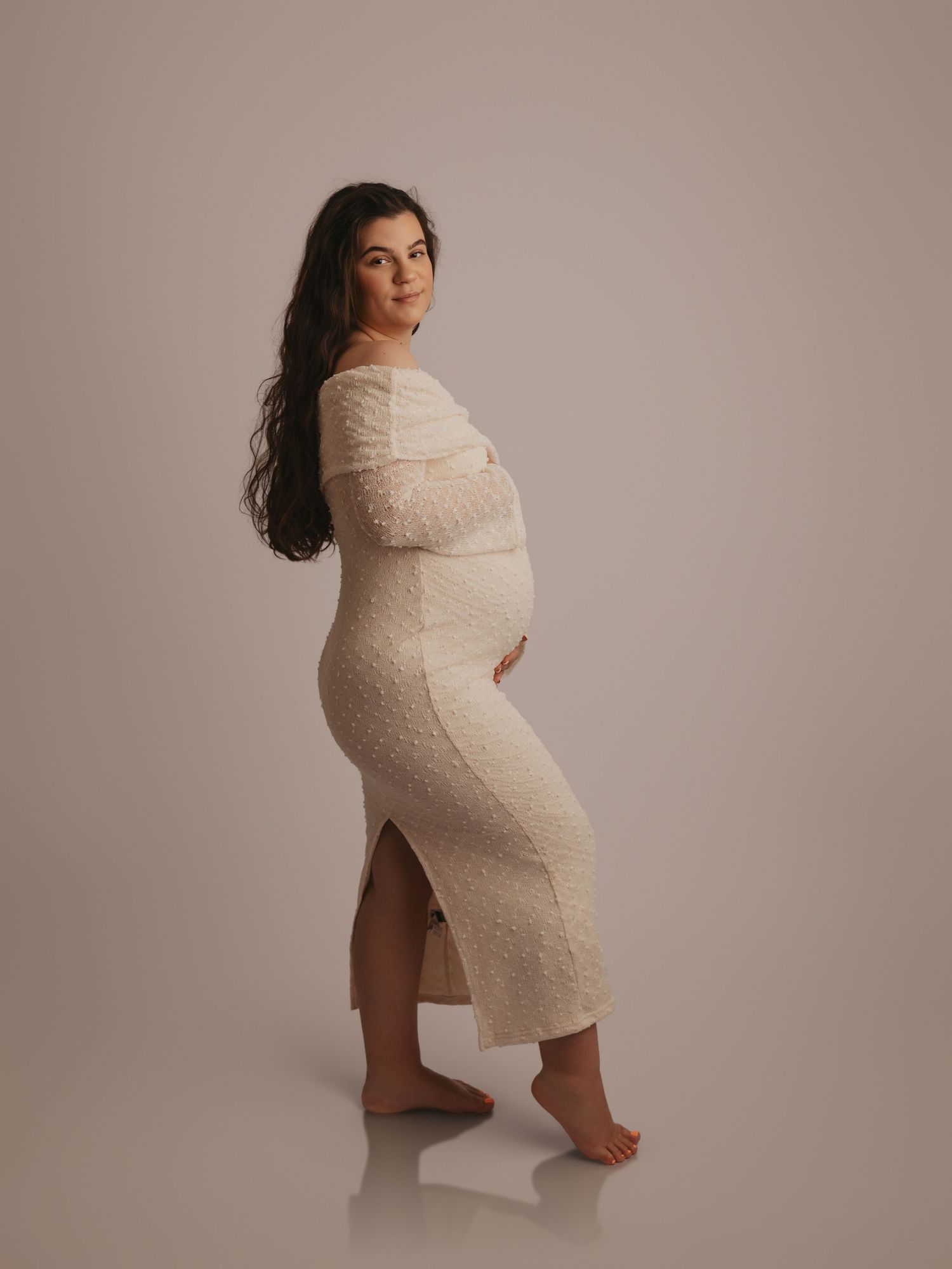 Pregnant woman in white lace off-shoulder maternity dress smiling, showing side slit in studio portrait.