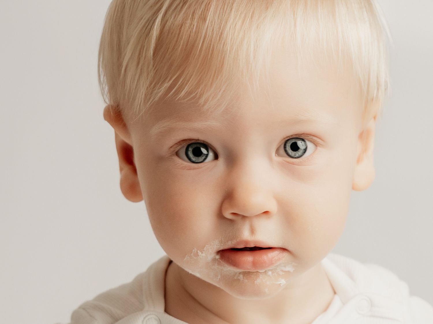 Close-up portrait of a blonde blue-eyed baby with white frosting on their chin during cake smash.
