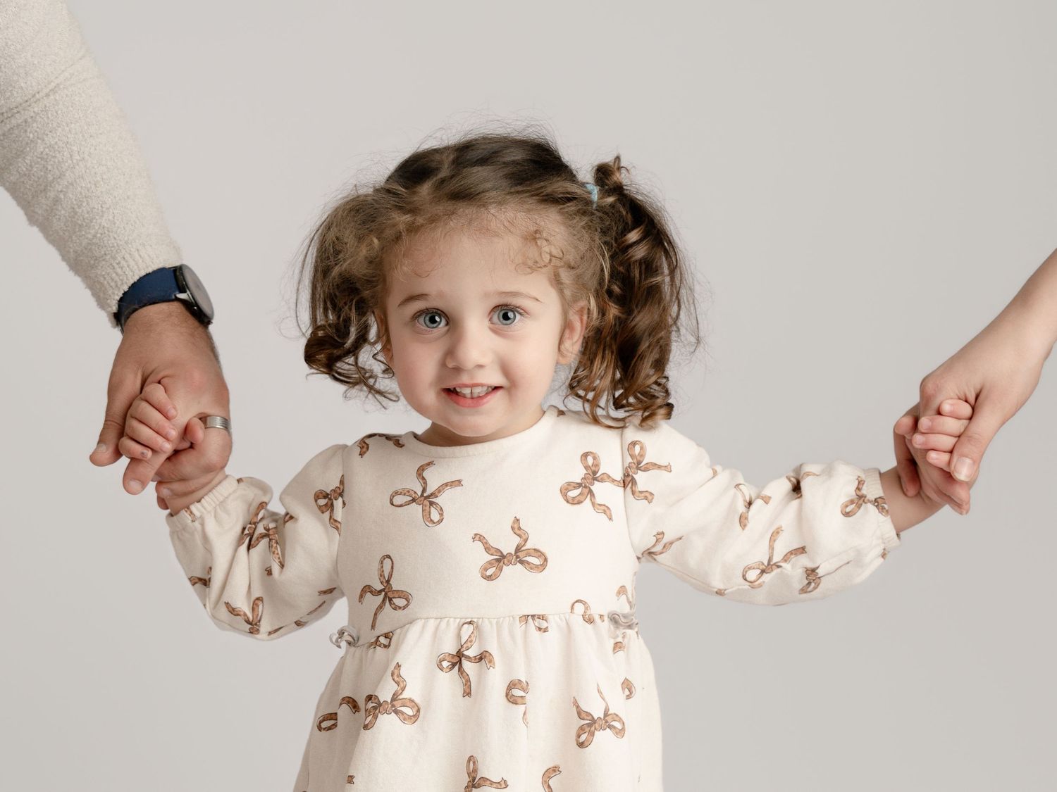 Happy toddler girl in bow-print dress smiling while holding both parents hands in studio.