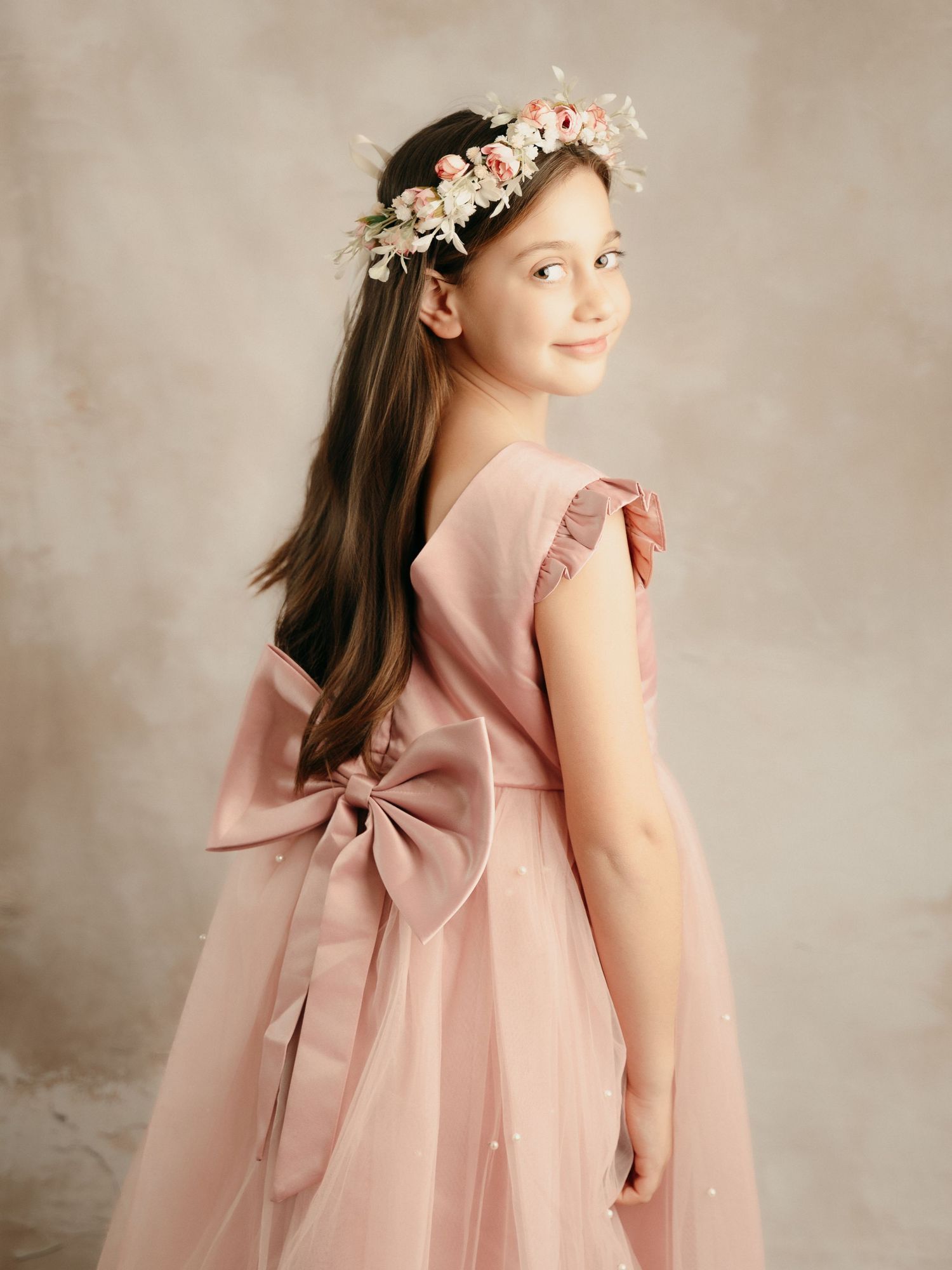 Young girl in pink tulle dress with large bow and floral crown, posing gracefully in studio portrait.