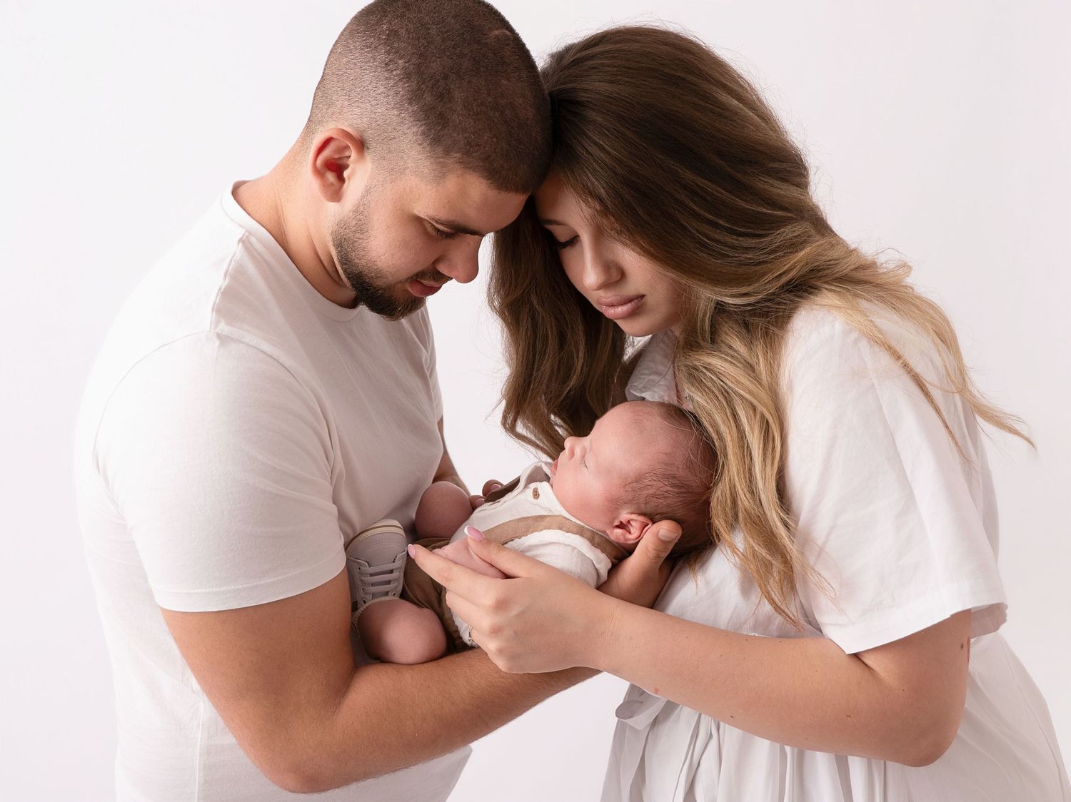 New parents in white outfits tenderly hold their sleeping newborn baby together in a studio portrait.