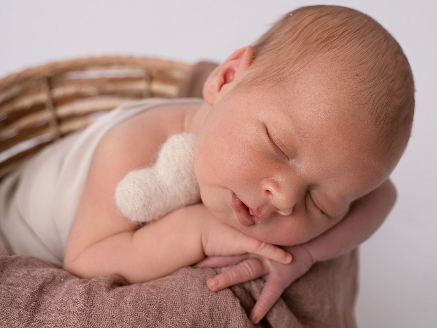 Sleeping newborn baby in a wicker basket with a small white teddy bear, wrapped in soft brown fabric.