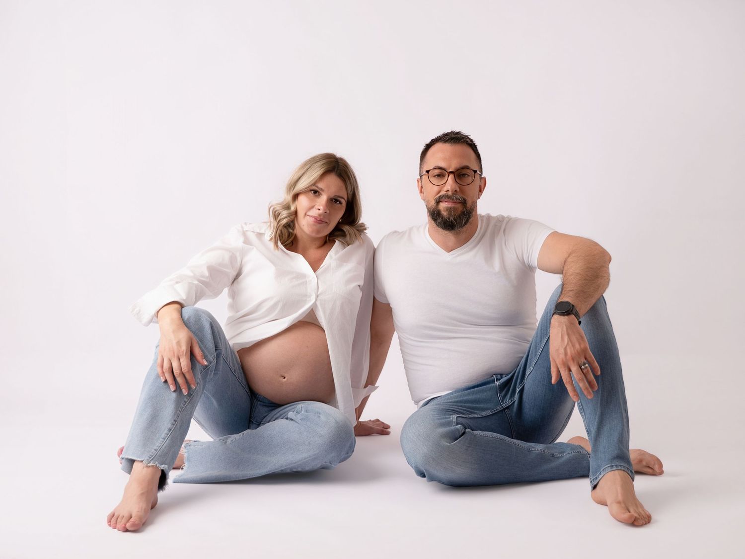 Pregnant woman and man sitting together on white background wearing white shirts and jeans in a maternity photo shoot.