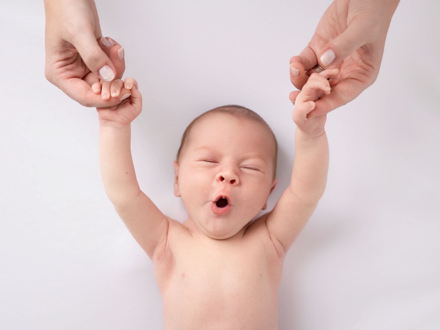 Newborn baby yawning with arms raised, held by adult hands on a white background.