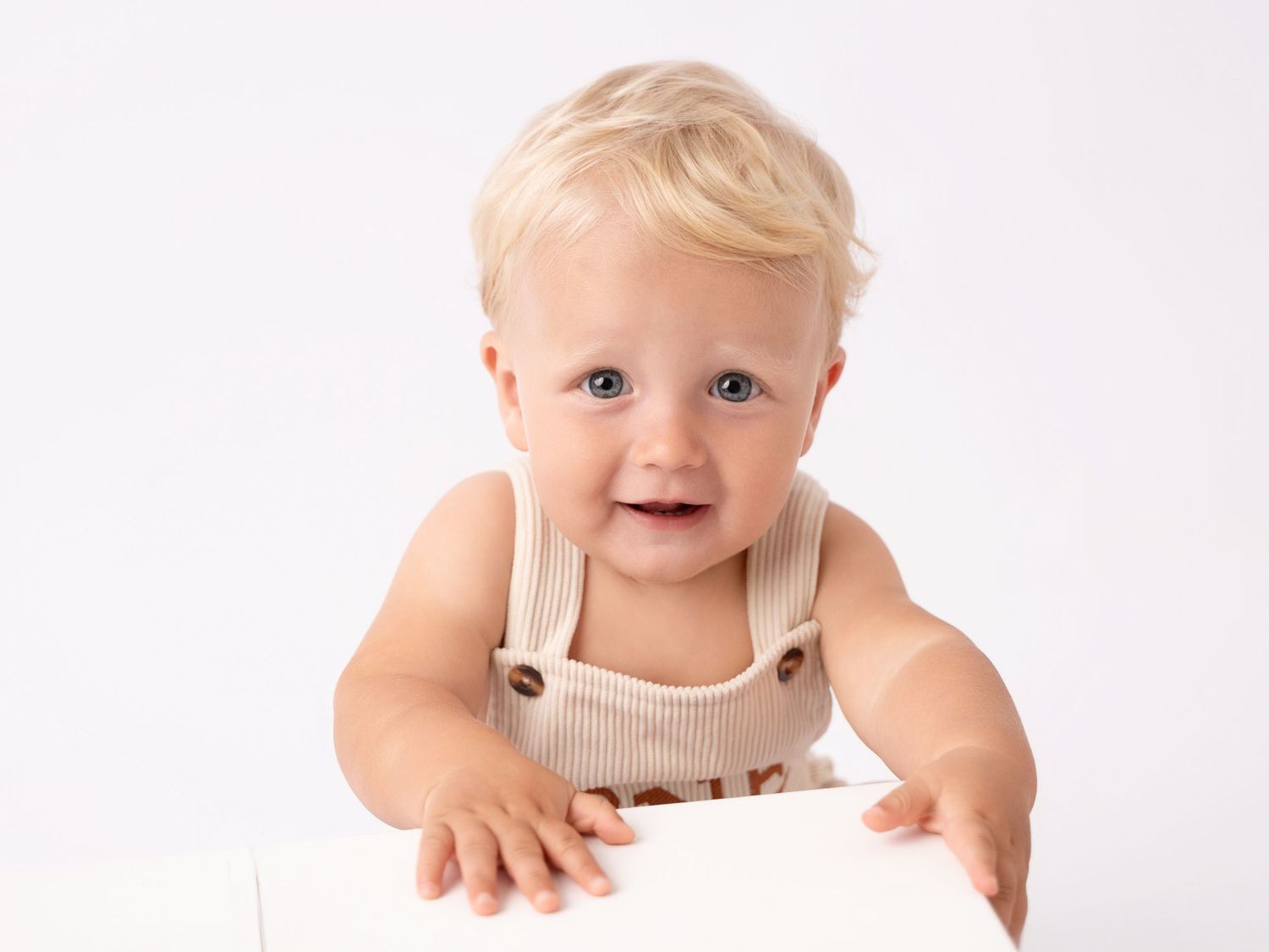 Smiling baby with blonde hair wearing a beige ribbed romper, posing against a white background.