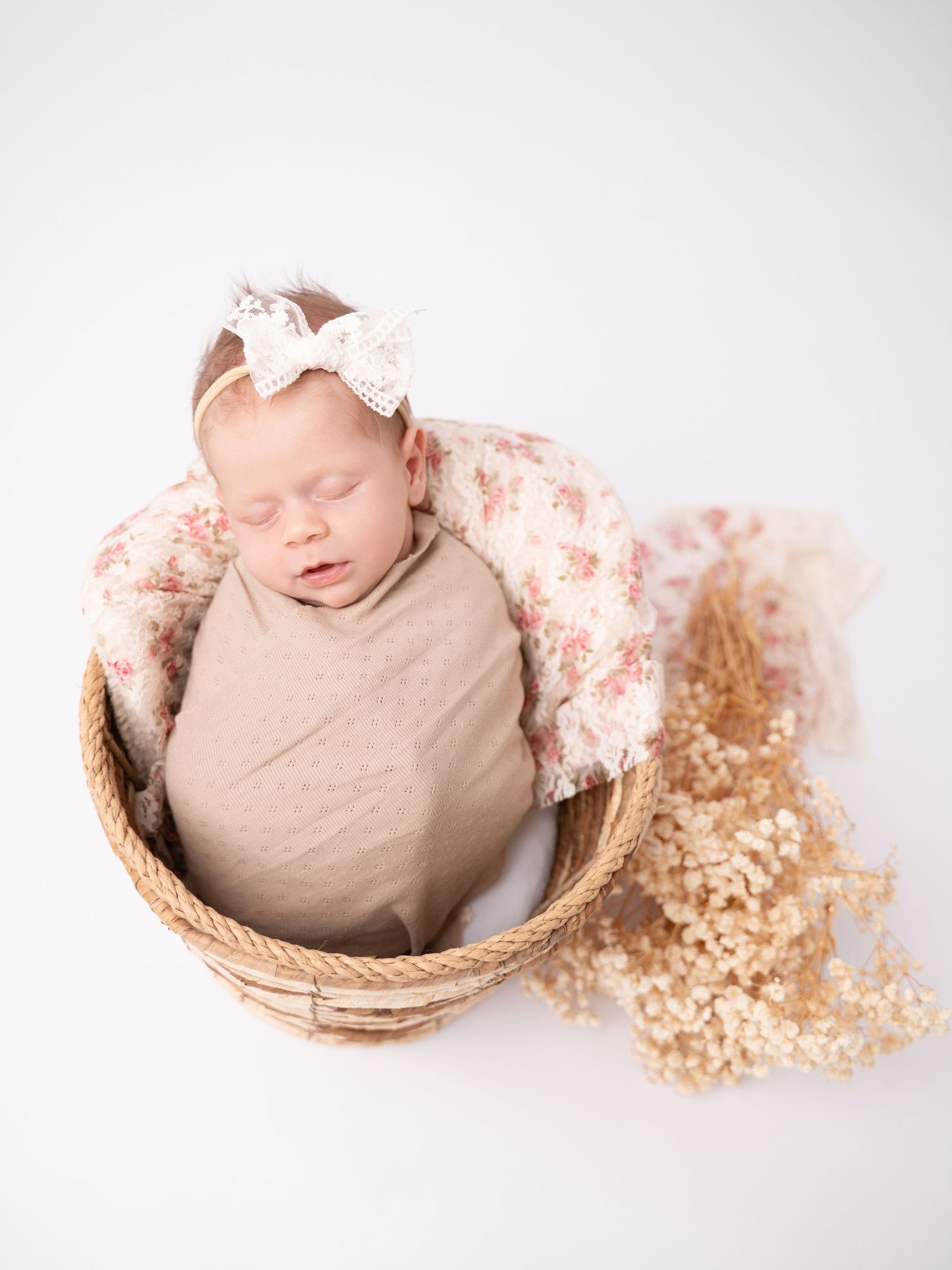 Newborn baby girl sleeping in a wicker basket, wrapped in beige, with a floral blanket and bow headband.