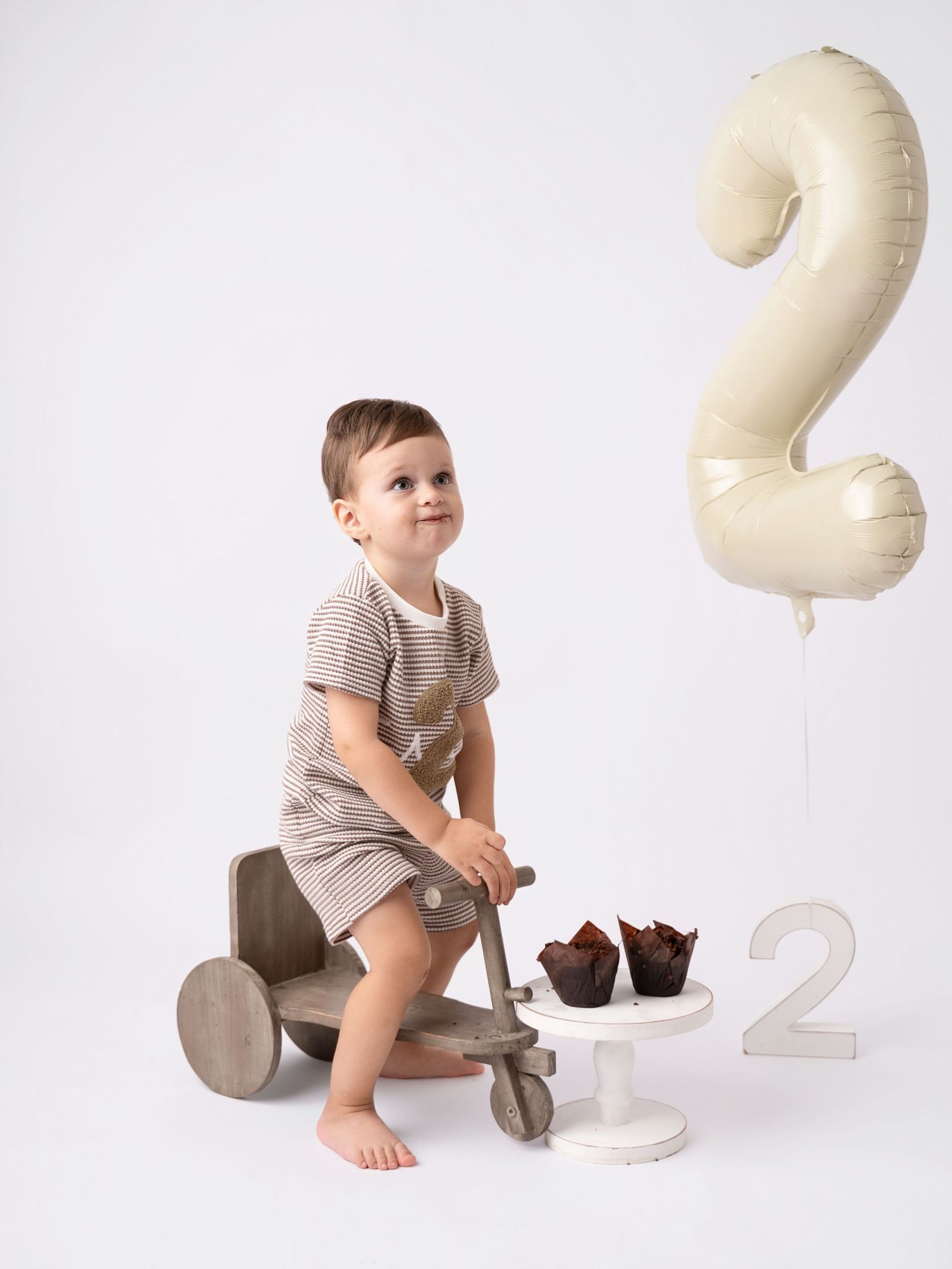 Happy toddler boy on wooden toy scooter beside number 2 balloon and muffins during birthday photo shoot.