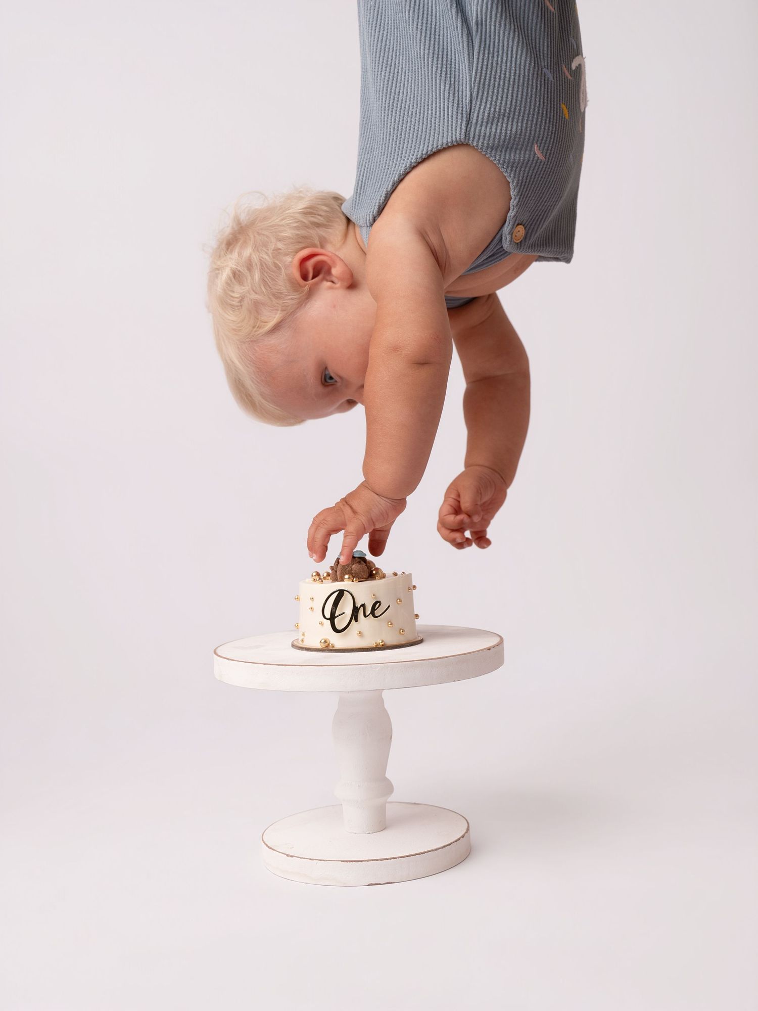 Toddler in blue romper reaching down to touch a white cake that reads One on a pedestal stand.