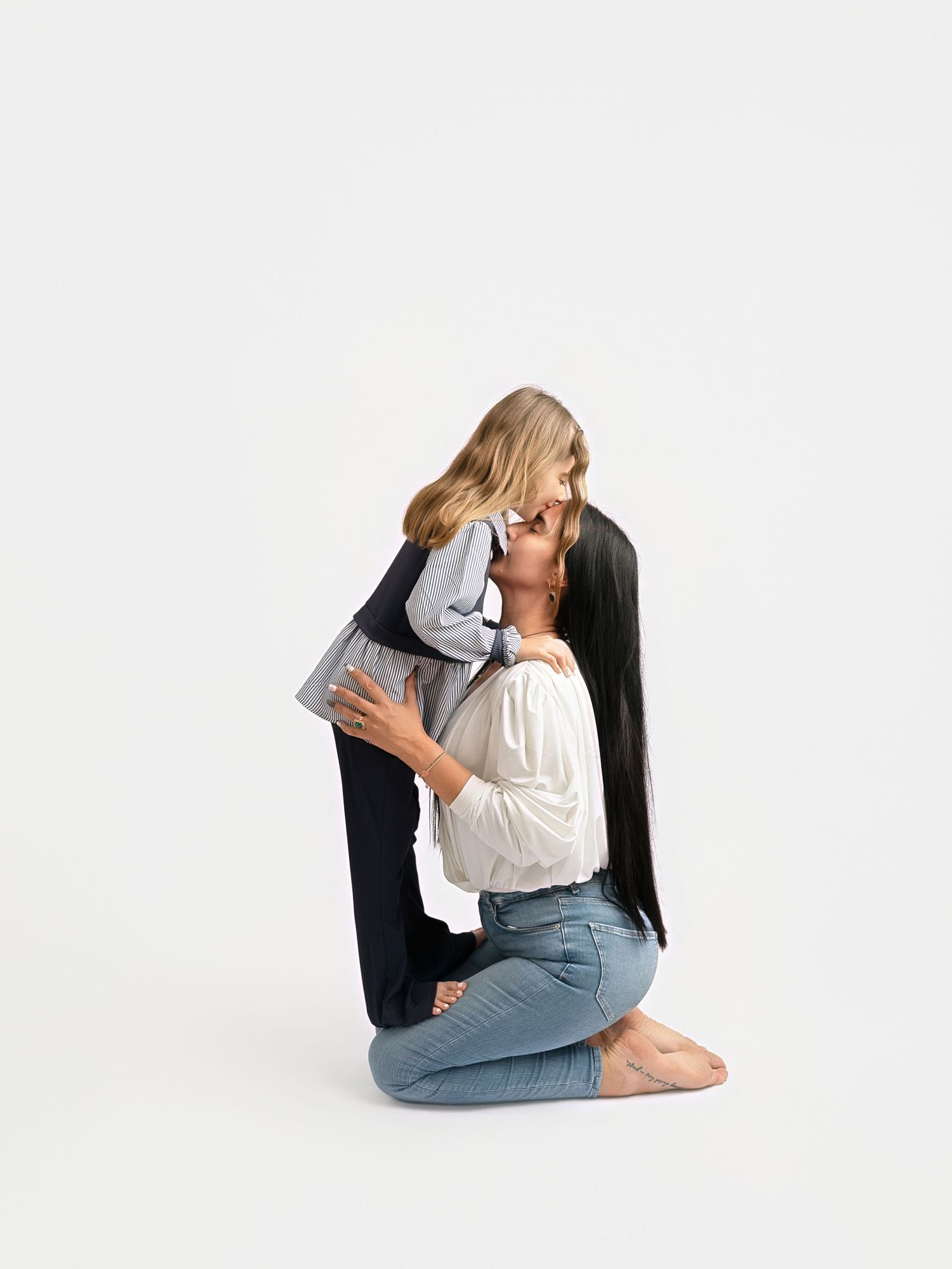A mother kneeling on the floor kissing her young daughter, both dressed casually in jeans and tops.