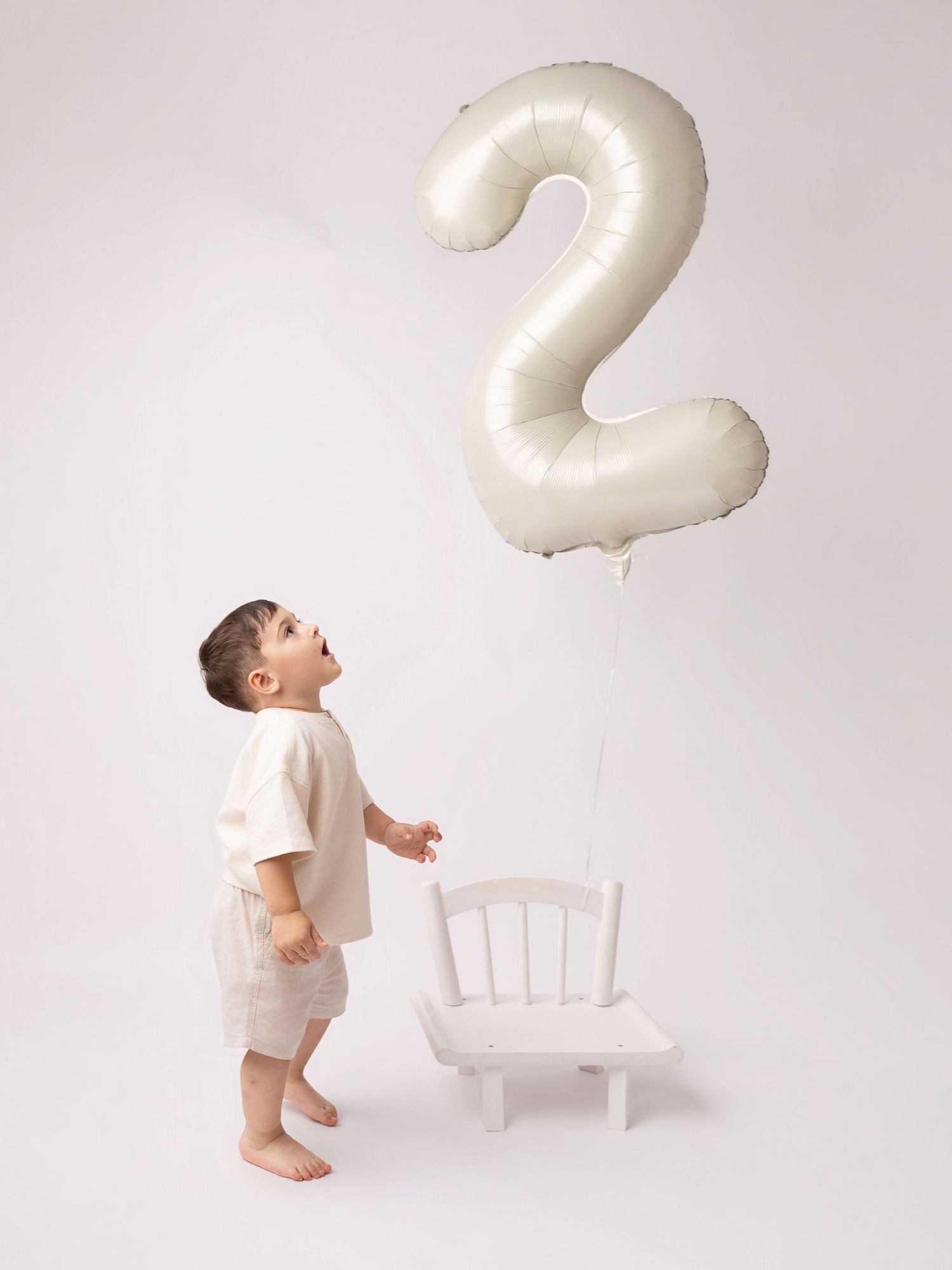 Toddler boy in beige outfit looking up at a number 2 foil balloon beside a small white chair.