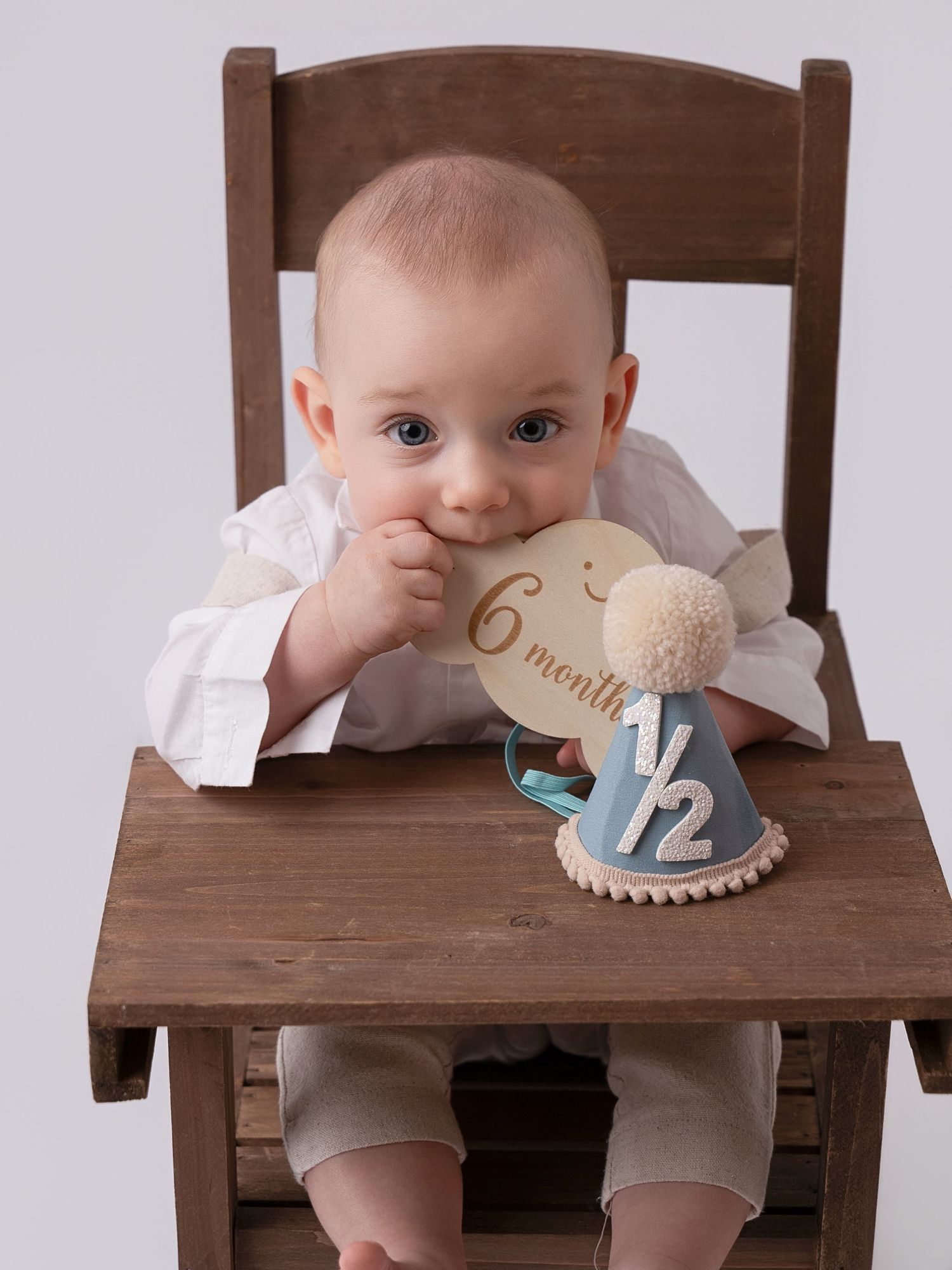 A baby sits in a wooden high chair chewing on a 6-month milestone disc with a half birthday hat nearby.