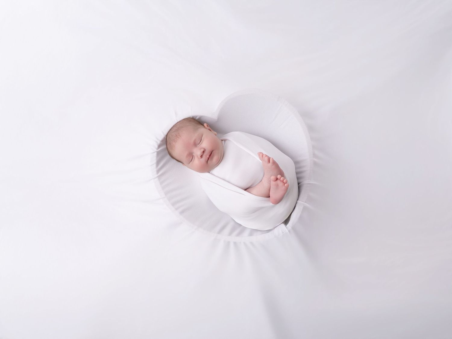 Sleeping newborn baby swaddled in white wrap, resting peacefully on a white heart-shaped pillow.