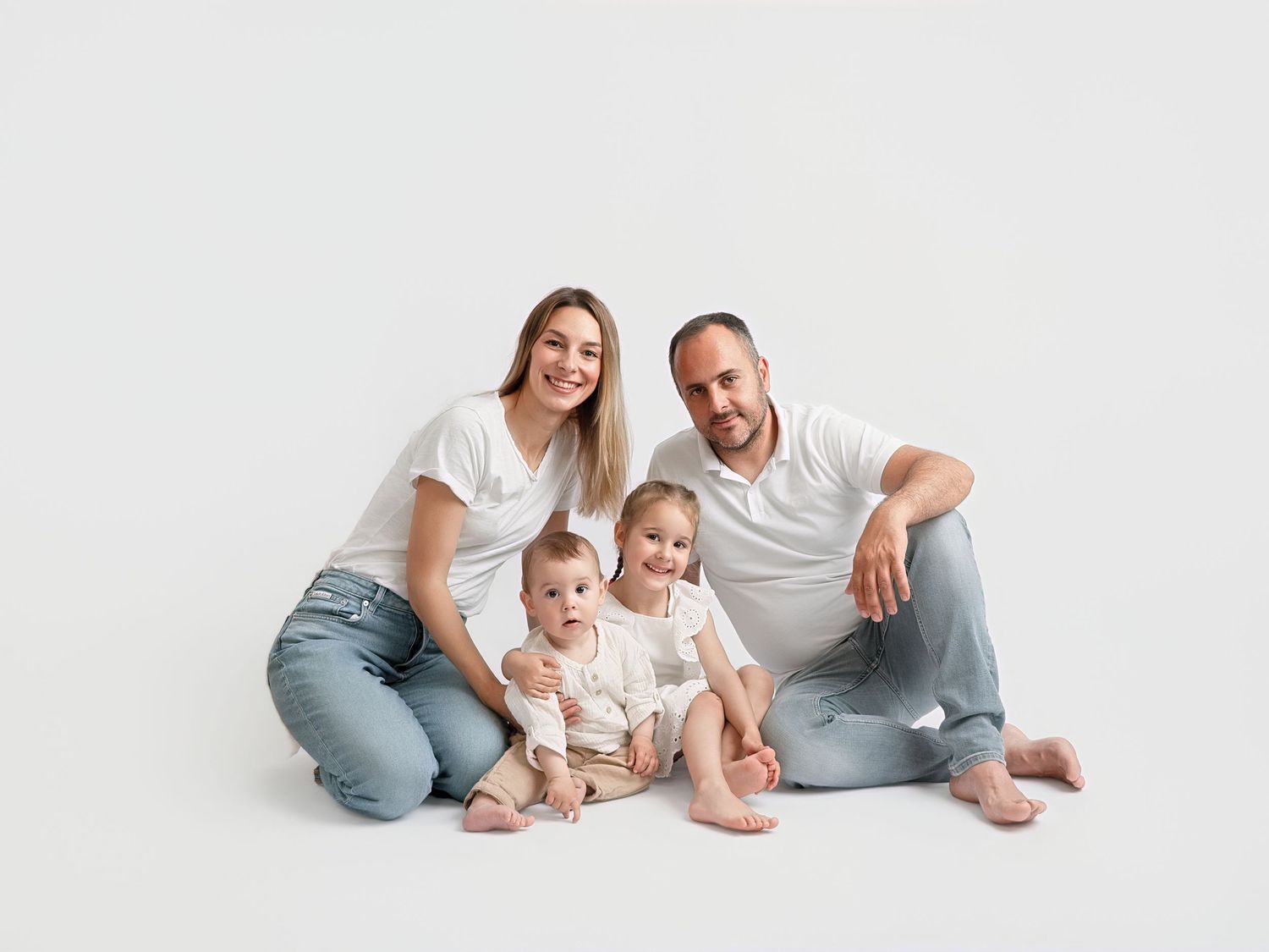 Happy family of four in white tops and jeans sitting together on white background for a studio portrait.