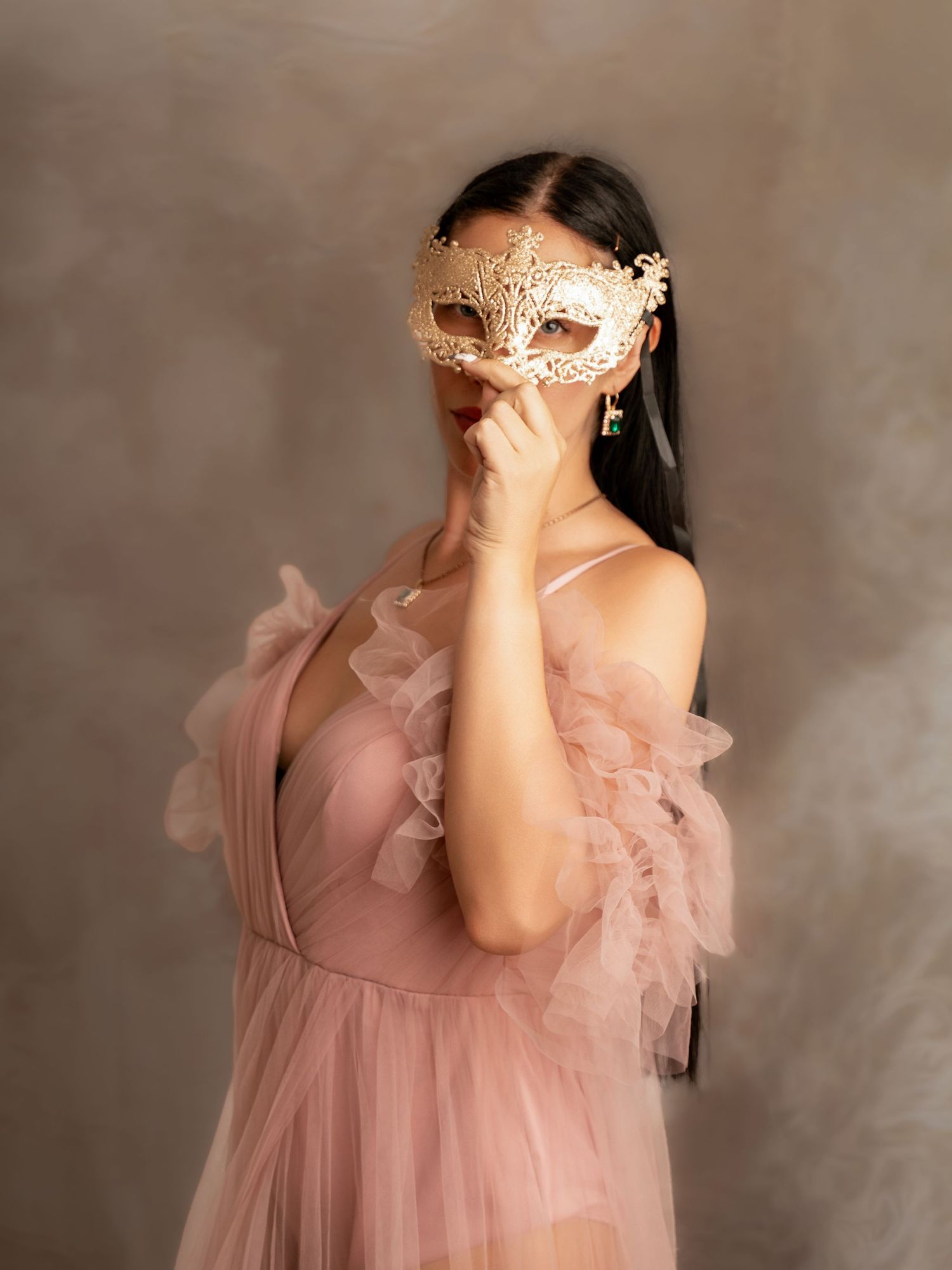 Woman in pink tulle gown holding ornate gold masquerade mask, posing elegantly against neutral studio backdrop.