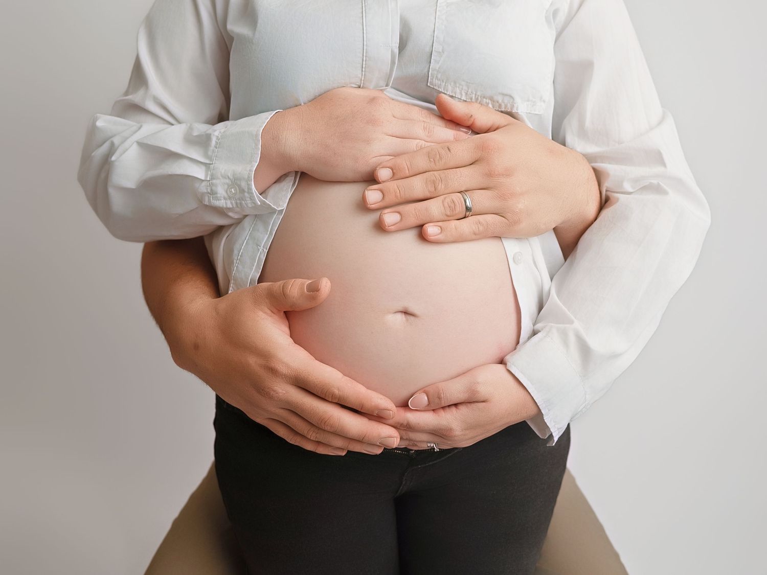 Pregnant woman in white shirt cradling her bare baby bump with both hands against a light background.