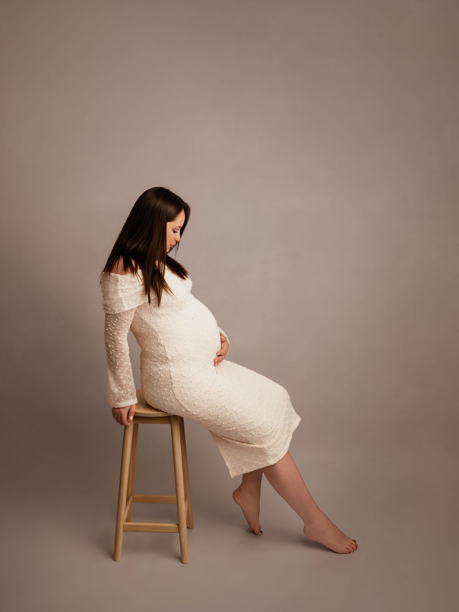 Pregnant woman in white lace dress sitting on wooden stool, cradling baby bump in professional maternity studio photo.