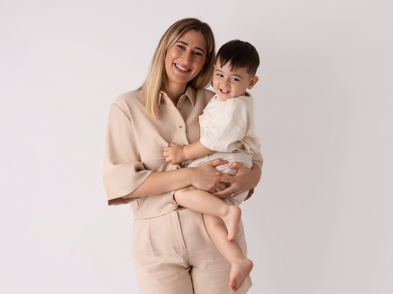 Smiling woman in beige outfit holding a happy toddler boy against a white studio background.