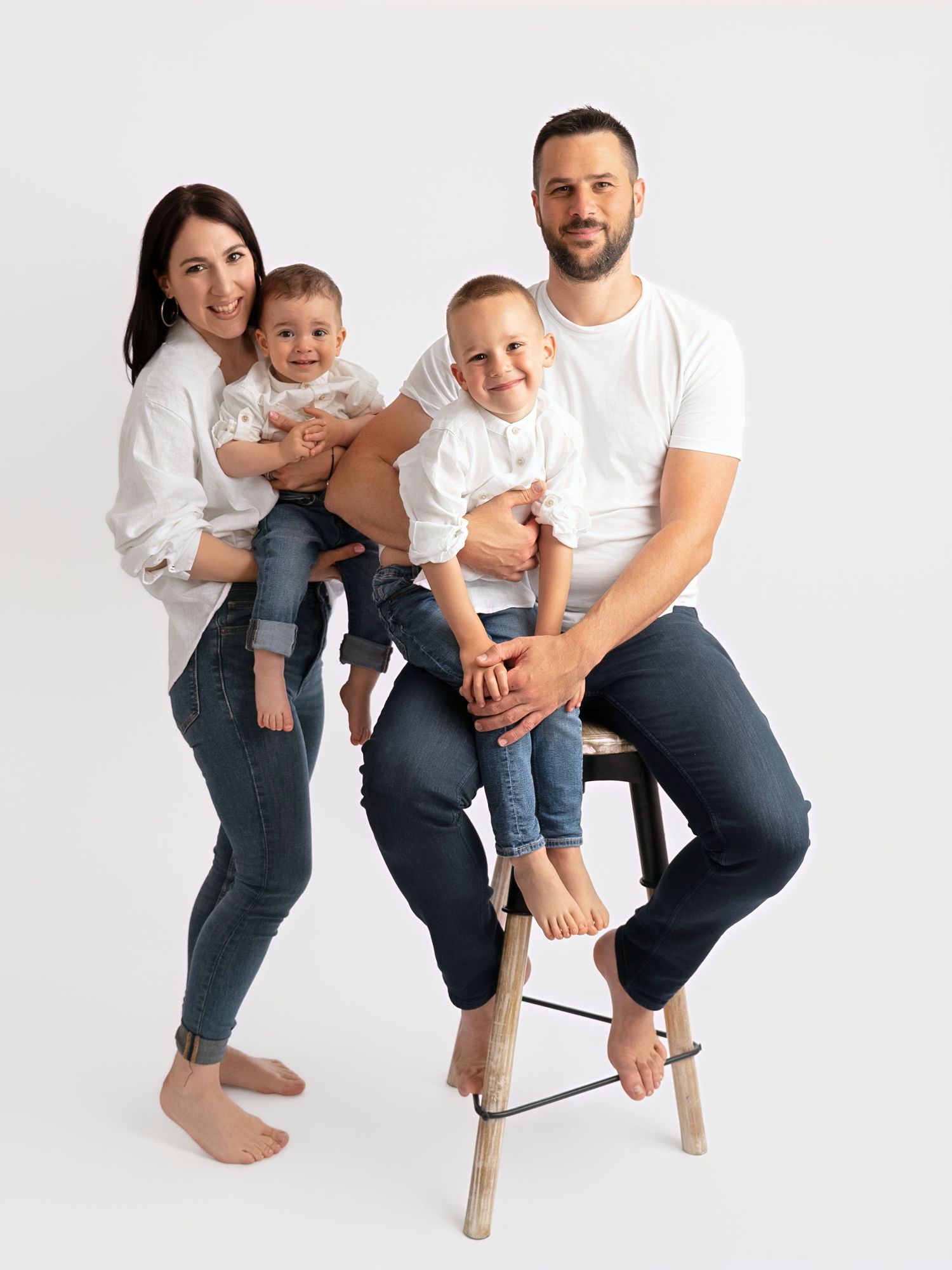 Happy family of four in matching white tops and jeans posing together on a stool against a white background.